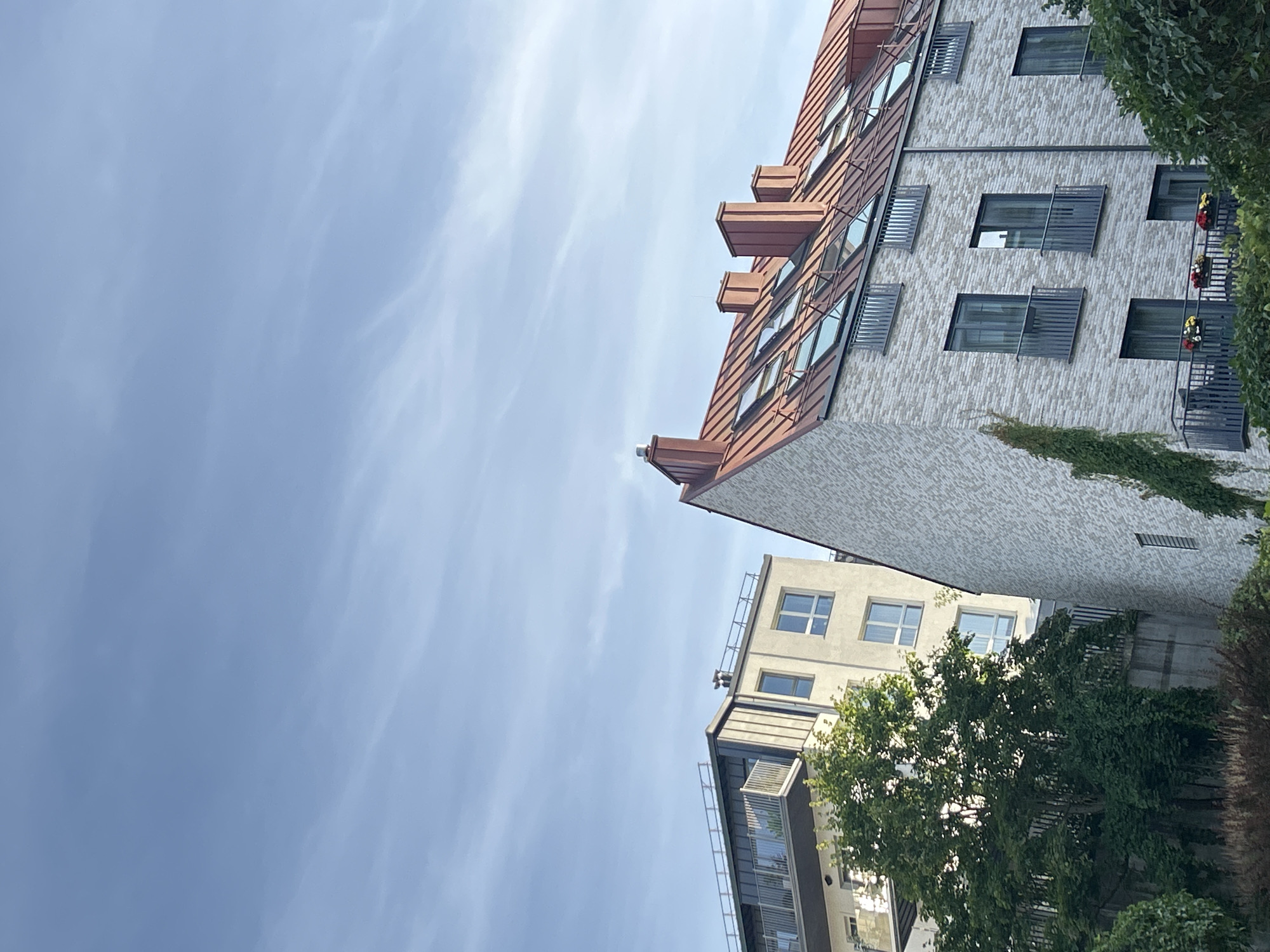 The image shows an outdoor scene of buildings against a blue sky with wispy clouds. The prominent building in the foreground is a multi-story structure with a light-colored brick facade and a rust-colored metal roof featuring multiple skylights and dormer windows. Several chimneys protrude from the roof. Balconies with dark railings are visible on the upper floors. To the left of this building, a taller, lighter-colored building with a dark, sloping roof and balconies is partially visible. Lush green trees and foliage obscure the lower portion of the buildings and the ground. The overall impression is one of modern architecture within an urban setting. This scene is located in the city of Vilnius, Lithuania.