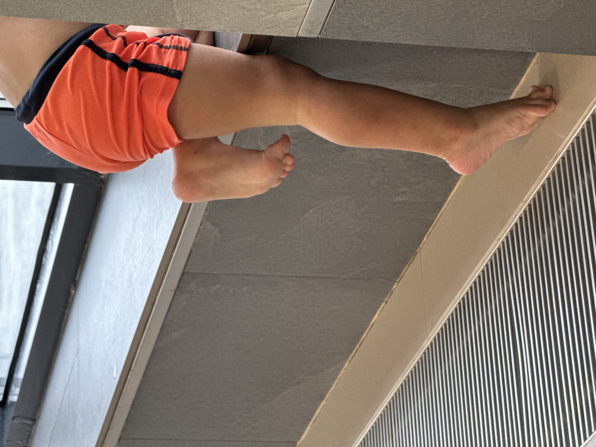 The image shows the lower body of a child wearing orange swim trunks with black trim. The child's left leg and foot are visible, with the foot poised on the edge of a surface. The child is leaning over, with their back to the camera. The environment appears to be a public pool or changing area, with grey tiled walls and a slatted metal drain cover. The lighting is natural, suggesting it is daytime. The image does not provide any information about the city of Chaikovsky, Russia.