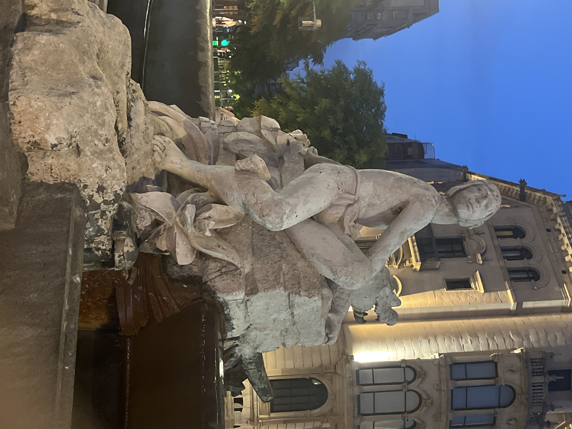 The image captures a daytime scene of a stone sculpture in Tarragona, Spain. The sculpture, possibly part of a fountain, depicts a human-like figure reclining on a rocky base. The figure is weathered, with details of its anatomy and drapery visible. In the background, traditional European architecture with arched windows and balconies lines a street. Trees with green foliage are interspersed between the buildings, suggesting an urban park or boulevard setting. The sky is a clear, light blue. The overall impression is one of a public art installation within a historic city.