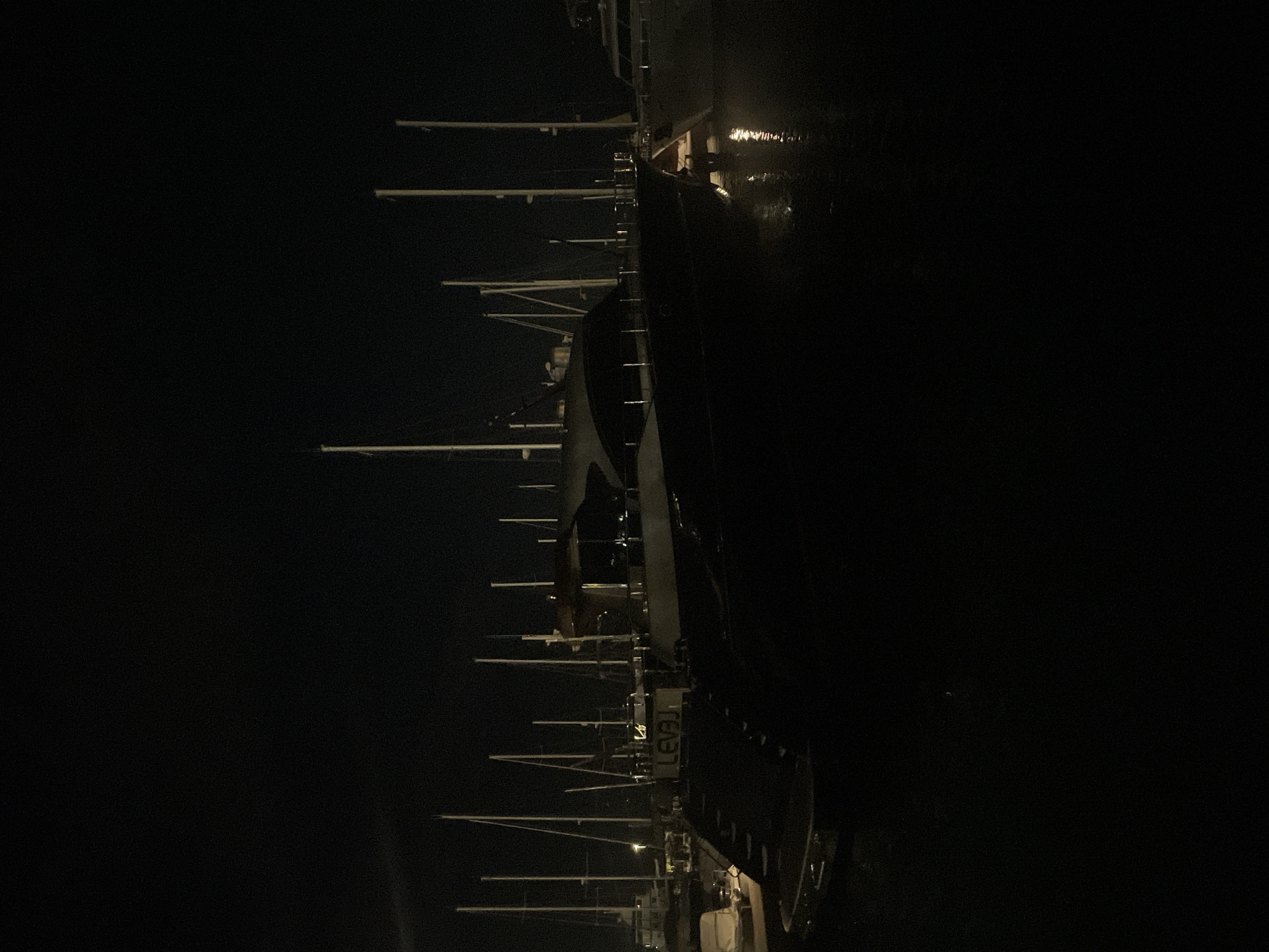 The image shows a nighttime scene at a marina in Bar, Montenegro. Several yachts and sailboats are docked, with their masts extending upwards against a dark sky. The primary focus is on a large, dark-colored yacht with "LEVAI" visible on its stern. The water reflects the lights from the boats, creating shimmering trails. The overall impression is one of a tranquil harbor at night.