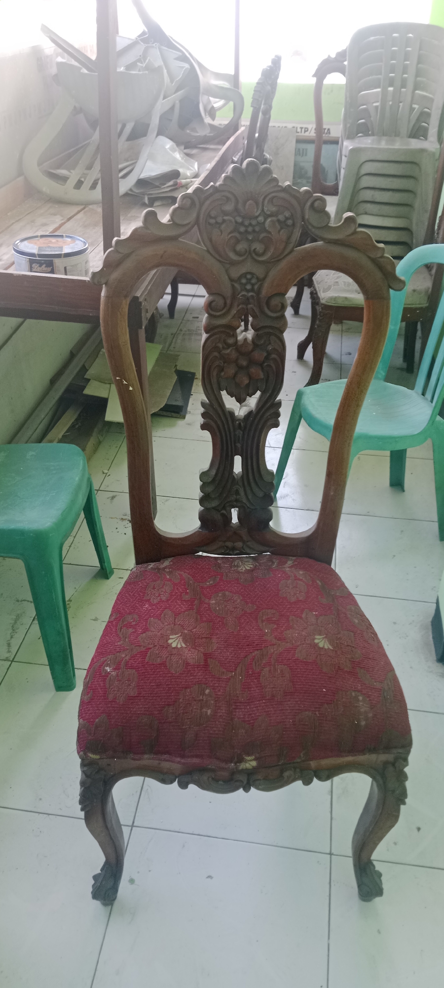 The image shows an ornate wooden chair with a red upholstered seat, positioned in a room with a tiled floor. The chair has intricate carvings on its back and legs. In the background, several plastic chairs are stacked, and some disassembled furniture parts are visible. The overall impression is of a storage area or a space undergoing renovation. The location is identified as Tuban, Indonesia, suggesting an indoor setting within a residential or commercial property in that city. There are no people or animals visible in the image.