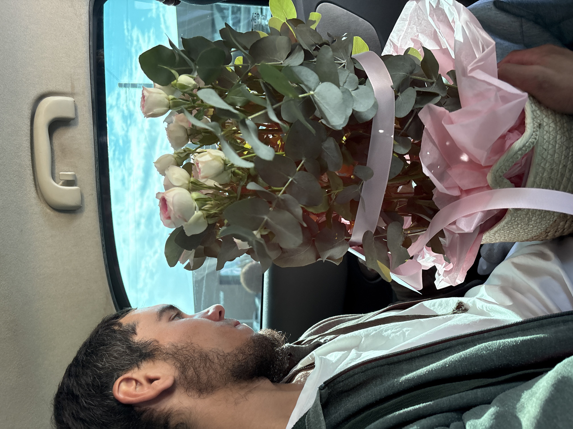 The image depicts a young man with dark hair and a beard looking out of a car window. He is wearing a white shirt and a grey jacket. In his lap, he holds a bouquet of light pink and white roses with green eucalyptus leaves, wrapped in pink tissue paper and tied with a pink ribbon. The bouquet is contained within a woven basket. The scene appears to be inside a vehicle, likely a car, with a portion of the car's interior, including a ceiling handle and a window showing a bright sky with clouds, visible. The photograph was taken in Pavlodar, Kazakhstan.