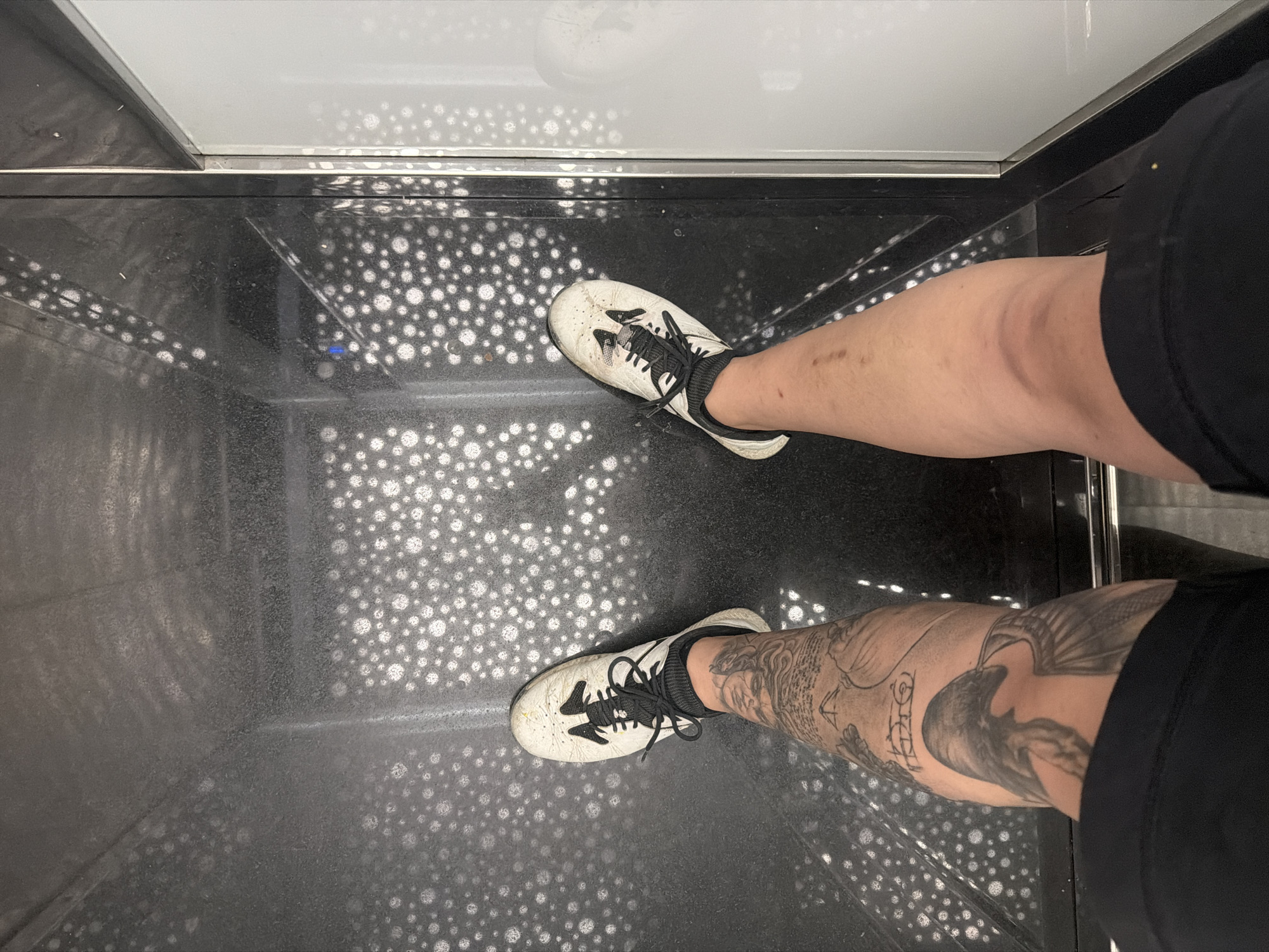 A first-person perspective shot shows a person standing in what appears to be an elevator. The person is wearing white athletic shoes with black laces and black socks. The left leg has extensive black ink tattoos of a bird, possibly a raven, and other designs. The right leg is bare from the knee down and shows some marks on the skin. The floor of the elevator is dark with a repeating pattern of small, light-colored circles. The elevator walls are metallic and reflective. To the right, a section of a white panel is visible. The scene does not contain any information or context that would allow for the identification of the city of Dolno Ablanovo, Bulgaria.