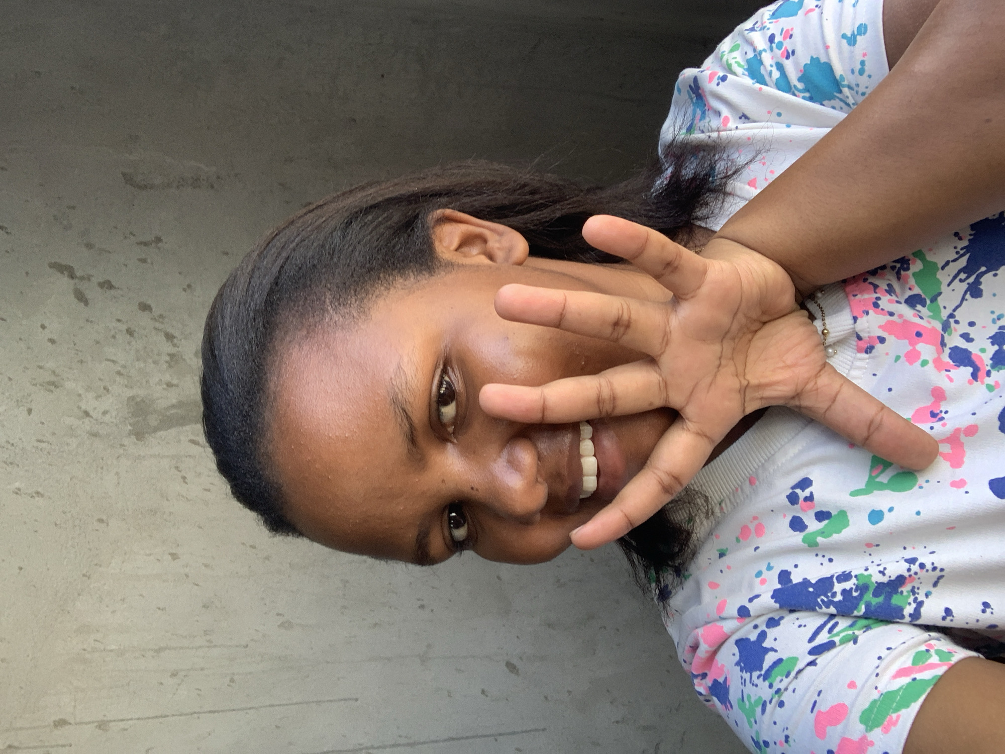A woman with dark hair and skin is smiling and holding up her left hand, partially obscuring her face. She is wearing a white t-shirt with splattered paint designs in blue, pink, and green. The background appears to be a concrete wall. It is not possible to determine the specific location in Dar es-Salaam, Tanzania from the image.