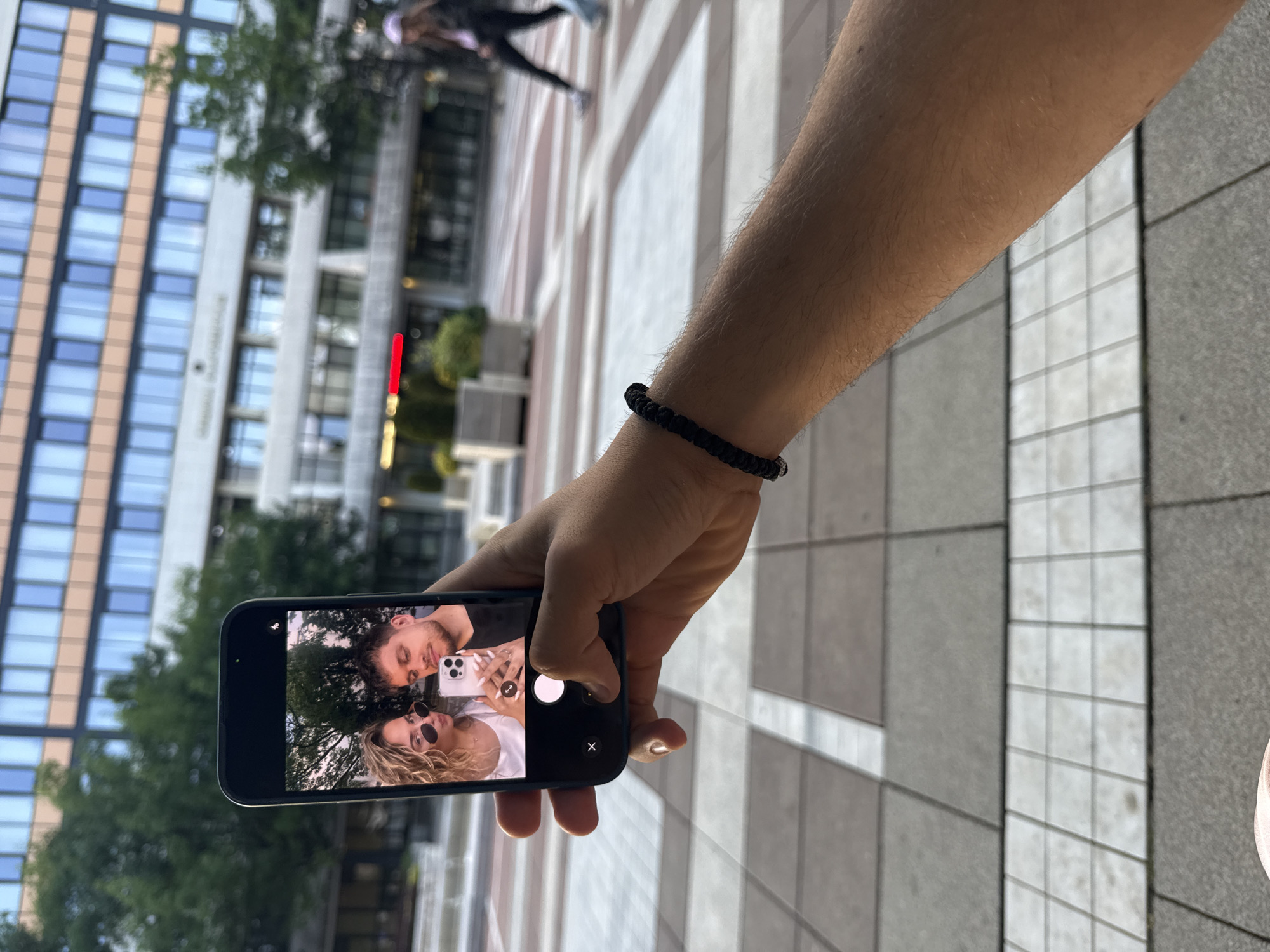 The image presents a first-person perspective of a person's arm holding a black smartphone horizontally, showcasing a selfie on its screen. The arm, lightly covered in hair, wears a black braided bracelet on the wrist. On the phone's display, two people are visible: a man with a beard looking towards the viewer, and a woman with blonde, curly hair and sunglasses, holding a white smartphone that reflects in her lenses. Lush green foliage appears in the background of the selfie.
The larger scene unfolds in an urban environment, specifically in Blagoevgrad, Bulgaria, indicated by the location context. Paved ground with grey and light-colored rectangular tiles stretches into the distance. Behind the hand and phone, modern, multi-story buildings with numerous windows and light-colored facades rise, some featuring vertical brown paneling. A blurred red vertical sign is faintly visible on one of the buildings. Trees are interspersed between the structures. In the far background, a blurred figure of a person is seen walking. The lighting suggests it is daytime, possibly late afternoon, with soft, clear light. The activity centers around viewing a selfie, potentially just taken or being reviewed. No discernible text is readable on the buildings or within the phone's interface, other than standard camera UI elements like a white circular shutter button and an 'X' symbol.