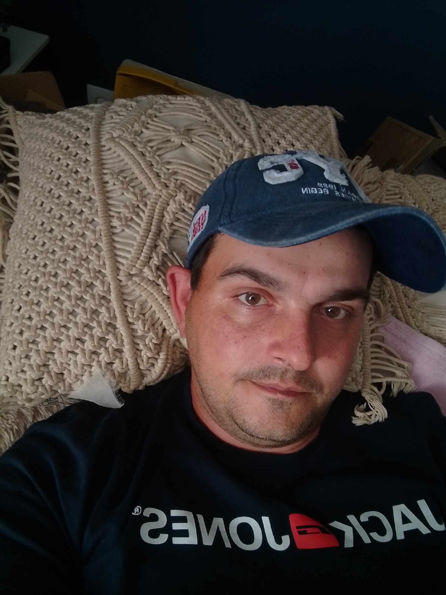 A man wearing a blue baseball cap and a dark blue sweatshirt with white text is lying down on a textured, cream-colored pillow. The text on his sweatshirt appears to be "JACK & JONES" reversed. He has short brown hair, brown eyes, and is looking towards the camera. The background is dark and out of focus, with some cardboard boxes visible. The image is taken from a close-up, head-and-shoulders perspective. No specific location in Wedel, Germany, is identifiable.