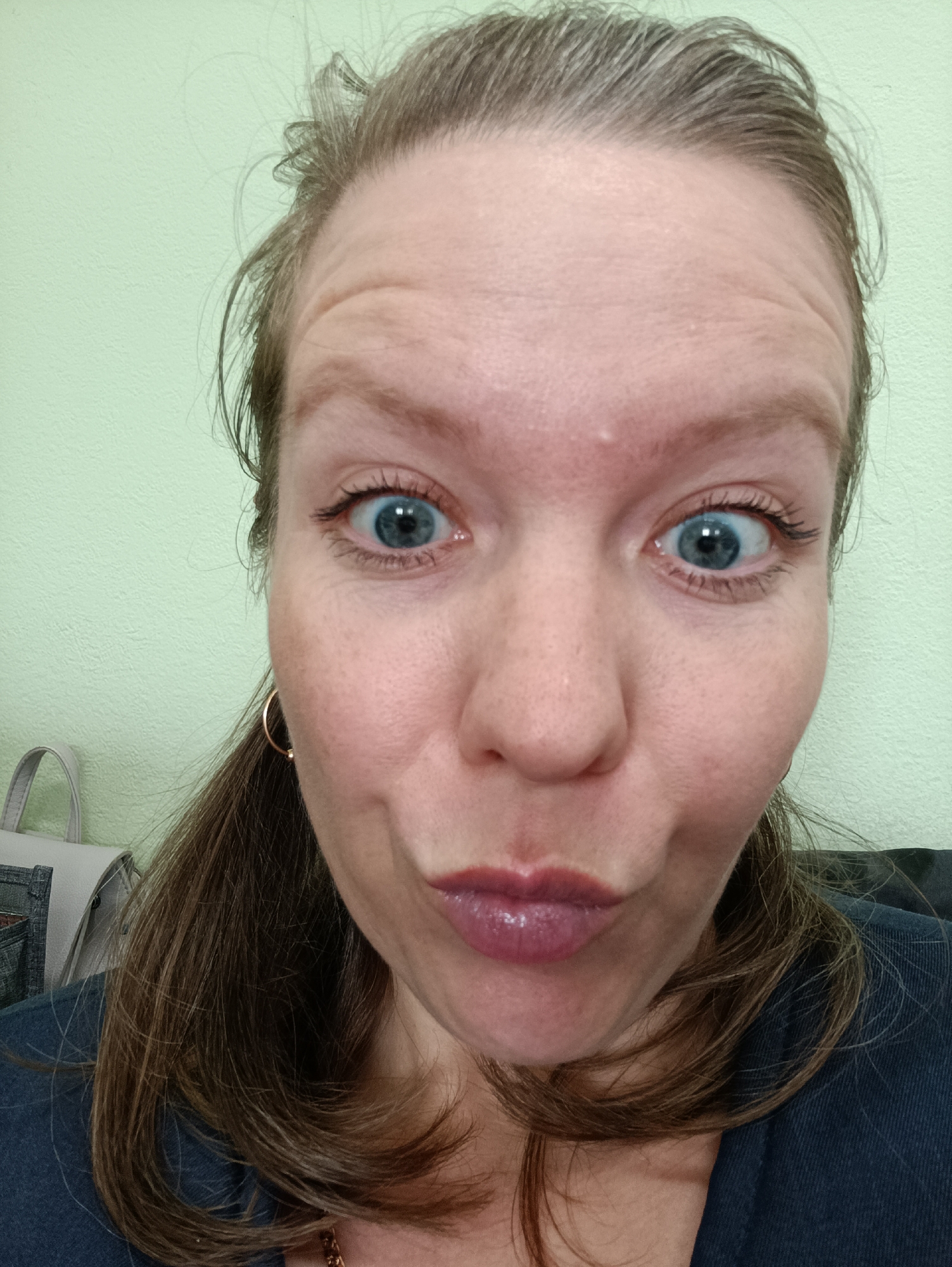 A close-up selfie depicts a woman with fair skin, blue eyes, and light brown hair pulled back loosely. Her expression is playful, with pursed lips and slightly widened eyes. She is wearing a dark blue shirt and a gold hoop earring is visible on her left ear. In the blurred background on the left, part of a gray bag with denim pockets can be seen. The wall behind her is a light green color. There is no information in the image to suggest that the photo was taken in the city of Chaikovsky, Russia.