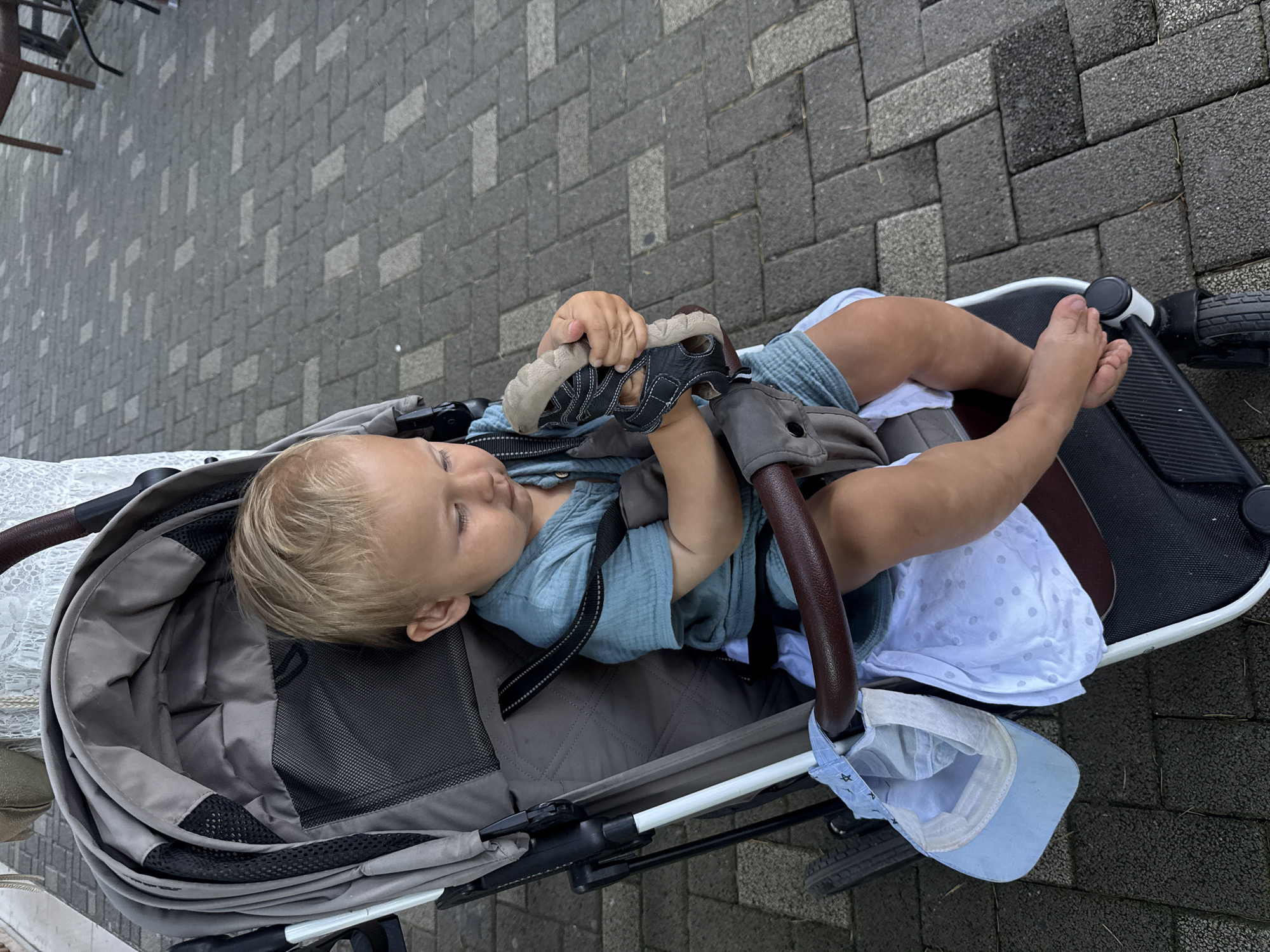 A blonde toddler with light skin, wearing a teal blue short-sleeved shirt and matching shorts, is seated in a gray stroller. The child is holding a black and tan sandal in their hands. A light blue baseball cap with a star pattern and a white cloth with light blue polka dots are attached to the stroller's handle. The stroller is positioned on a paved walkway made of gray and dark gray rectangular bricks, arranged in a herringbone pattern. The background shows a glimpse of a white lace tablecloth and brown chairs, suggesting an outdoor seating area, possibly a cafe or restaurant. The scene appears to be a sunny day, with the stroller's canopy open. The location is specified as Sochi, Russia.