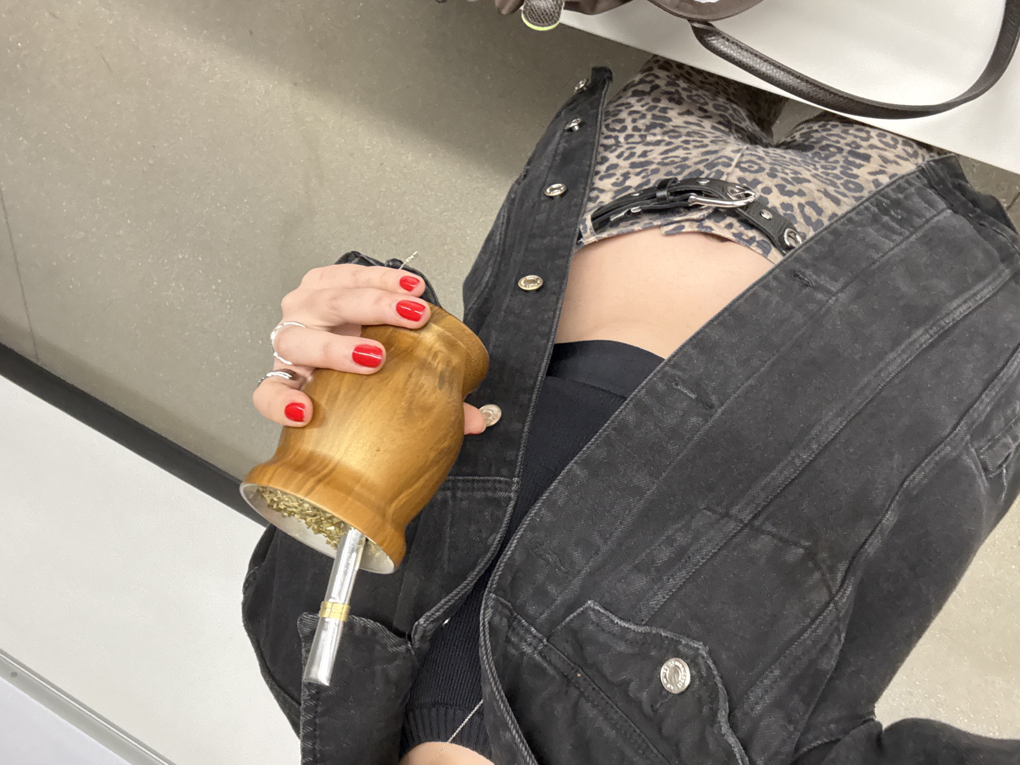 The image captures a close-up, slightly tilted view of a person's upper torso and hands, likely indoors. The perspective is looking down, with the person positioned diagonally across the frame.
The central subject is a hand, adorned with several silver rings and bright red nail polish, holding a light brown wooden mate gourd. The gourd is filled with yerba mate and contains a silver *bombilla* (drinking straw). This suggests the activity of drinking or preparing to drink mate.
The person is dressed in a black denim jacket, worn open to reveal a leopard print top or dress underneath, and a glimpse of a black bra or crop top. The jacket features silver buttons and visible stitching details. The person's midriff is partially visible between the jacket and the leopard print garment.
The background consists of a plain, light gray or beige surface, possibly a wall or counter, with a dark line indicating an edge. In the upper right corner, a dark brown leather strap, possibly from a bag, is visible. No specific time of day or weather conditions can be inferred, and the setting appears to be a simple indoor environment. Given the context of Mexico City, Mexico, this implies the cultural practice of drinking mate is present there. There is no visible text in the image.