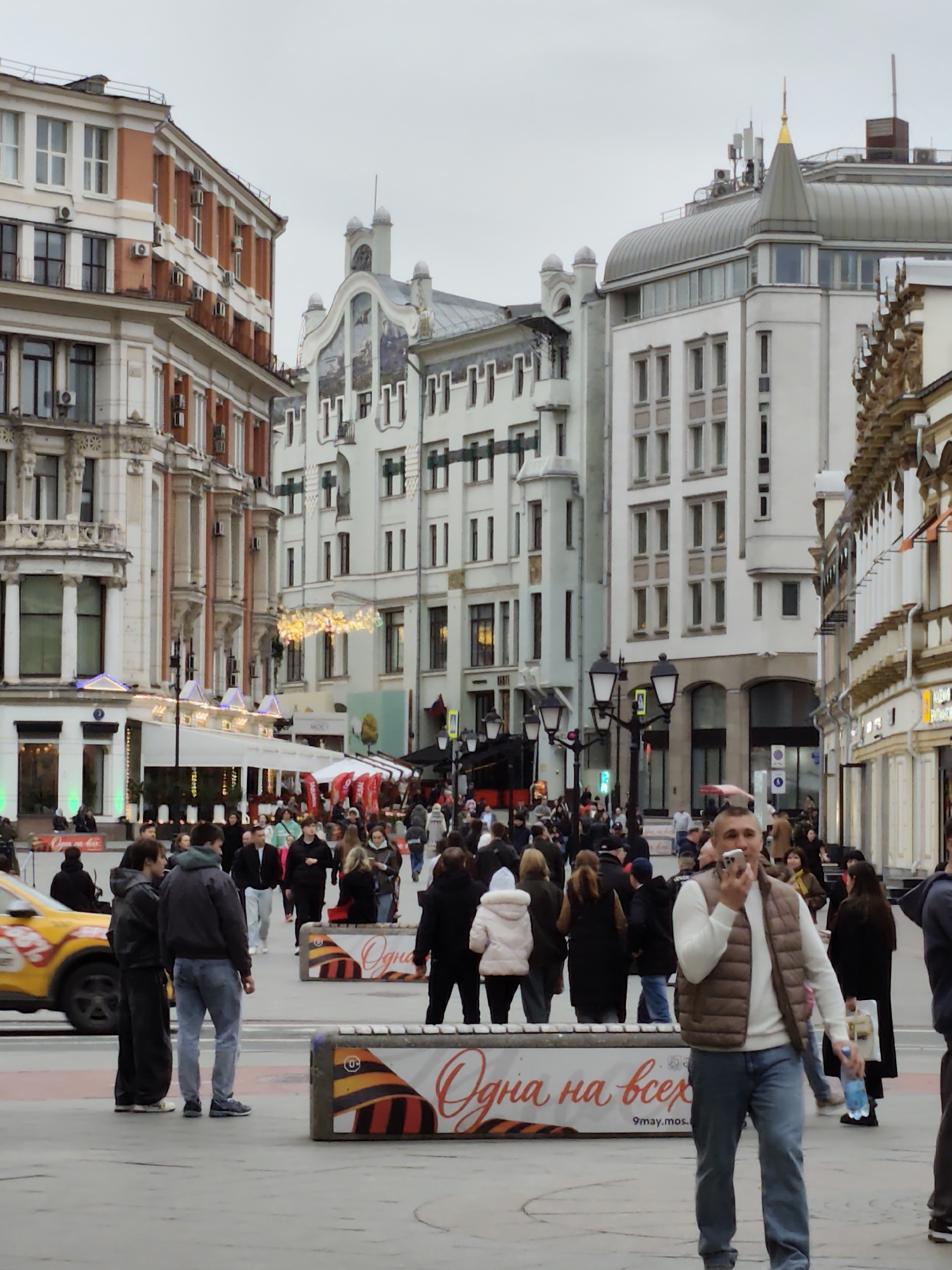 A street scene in Moscow, Russia, shows a crowded pedestrian area with a variety of buildings. The architectural styles range from ornate to modern. People are casually dressed, walking through the area. A man in the foreground holds a cell phone up to his mouth as if recording a voice memo or talking. Barriers with the phrase "Одна на всех" are present. A yellow taxi moves in the background. The weather appears overcast.