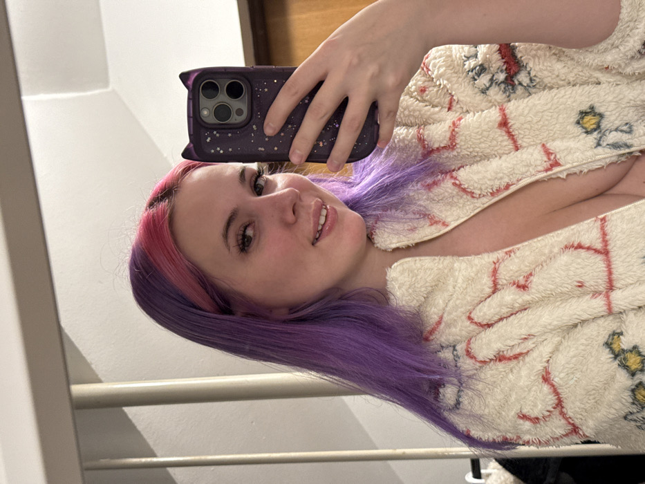 A young woman is taking a selfie in a mirror. Her hair is dyed pink at the roots and transitions to purple. She wears a patterned, plush robe. She holds a purple phone with a cat-ear case. The background shows a white wall and a glimpse of a wooden door.