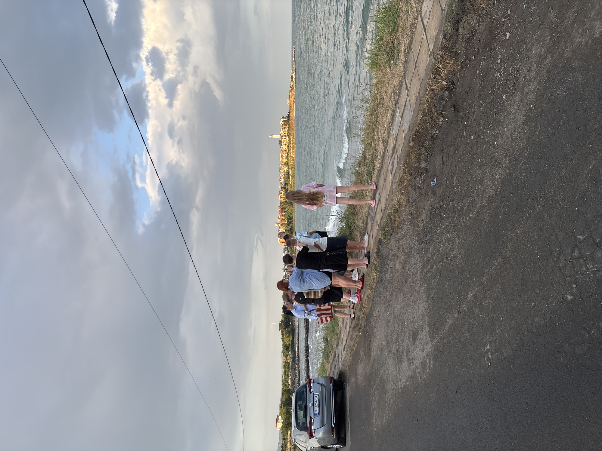 The image depicts a group of six people, including four children and two adults, gathered by the sea in Aheloy, Bulgaria. They are standing on a paved walkway next to a road, with a silver car parked to their left. The sea is visible in the background, with a distant town featuring a lighthouse and colorful buildings. The sky is cloudy, with some patches of blue and sunlight breaking through. The overall atmosphere appears to be casual and relaxed, with the group seemingly enjoying the coastal scenery.