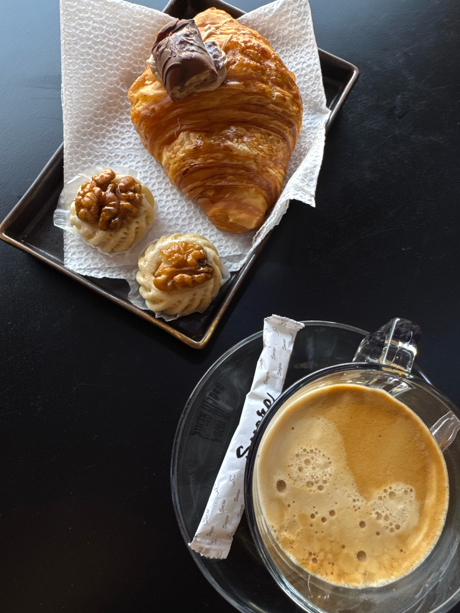 A close-up, top-down shot features a pastry and coffee on a dark, possibly black, textured surface, likely a table. The arrangement is on the right side of the frame. In the upper left quadrant, a dark metal tray holds a croissant topped with two pieces of chocolate. Beside the croissant are two small, intricately shaped cookies, each adorned with a walnut. The tray rests on a white, textured paper napkin. To the lower right, a clear glass mug filled with frothy coffee sits on a matching glass saucer. A single-serving packet of sugar, white with black text, lies diagonally across the saucer and next to the mug. The background is a solid, dark color, with subtle variations in tone suggesting a textured surface. There are no people visible in the image. The overall impression is one of a simple, appealing breakfast or snack. The scene is unlikely to be in Essaouira, Morocco, as the coffee packet clearly shows the word "Sucre," which is French for sugar, and the overall aesthetic does not strongly suggest a Moroccan setting.
