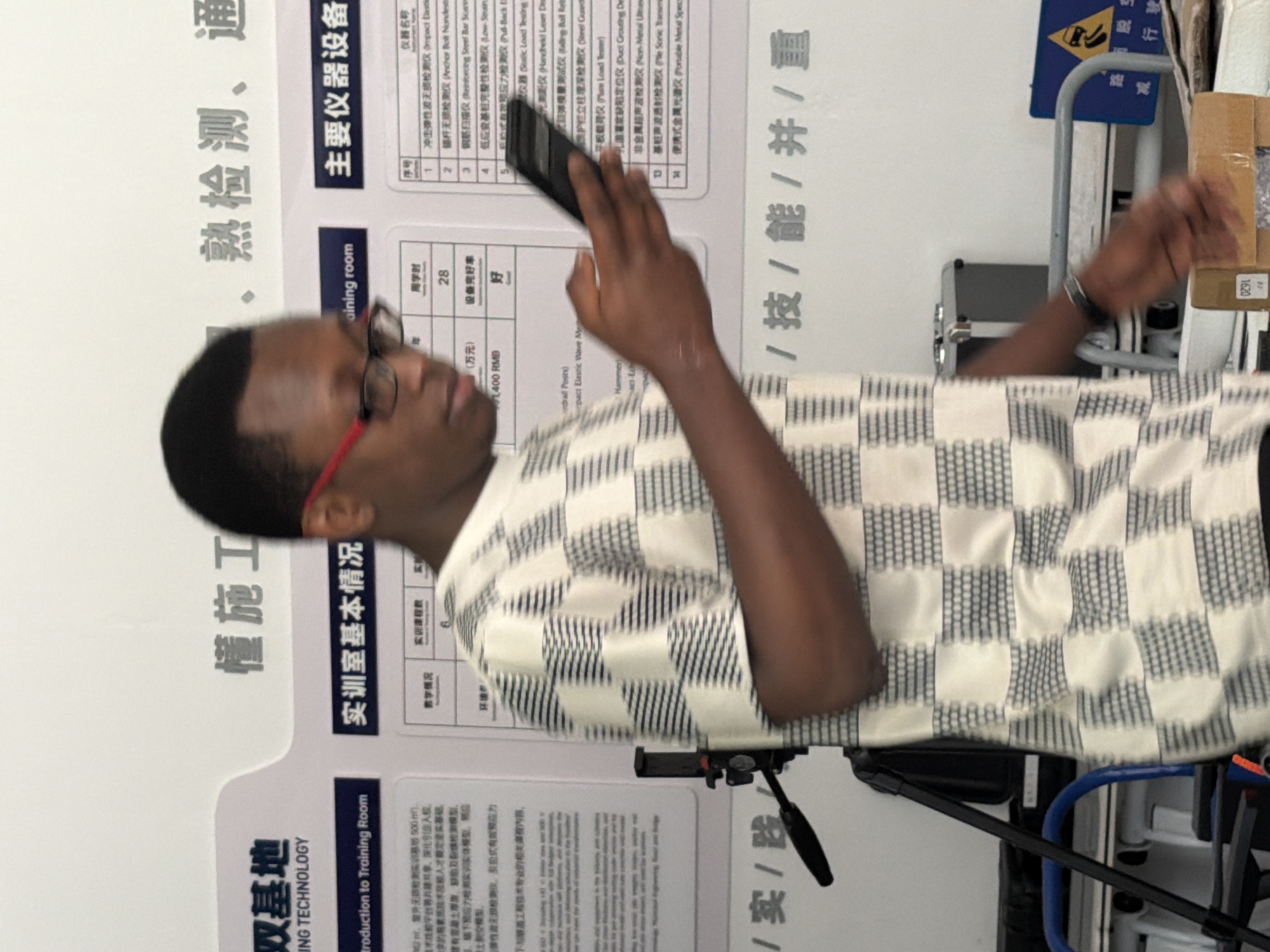 A young Black man wearing glasses and a checkered shirt holds a smartphone in his right hand, seemingly interacting with it. He is positioned in front of a white wall adorned with Chinese text and graphics. The text appears to be from a training or educational institution, with headings like "实训室基本情况" (Training Room Basic Information) and "主要仪器设备" (Main Instruments and Equipment). Several tables with chairs and equipment are visible in the background, suggesting a laboratory or workshop setting. The man's posture and the smartphone in his hand imply he is engaged in a task related to the displayed information or a personal activity. The overall environment suggests an indoor space associated with technical training or research.