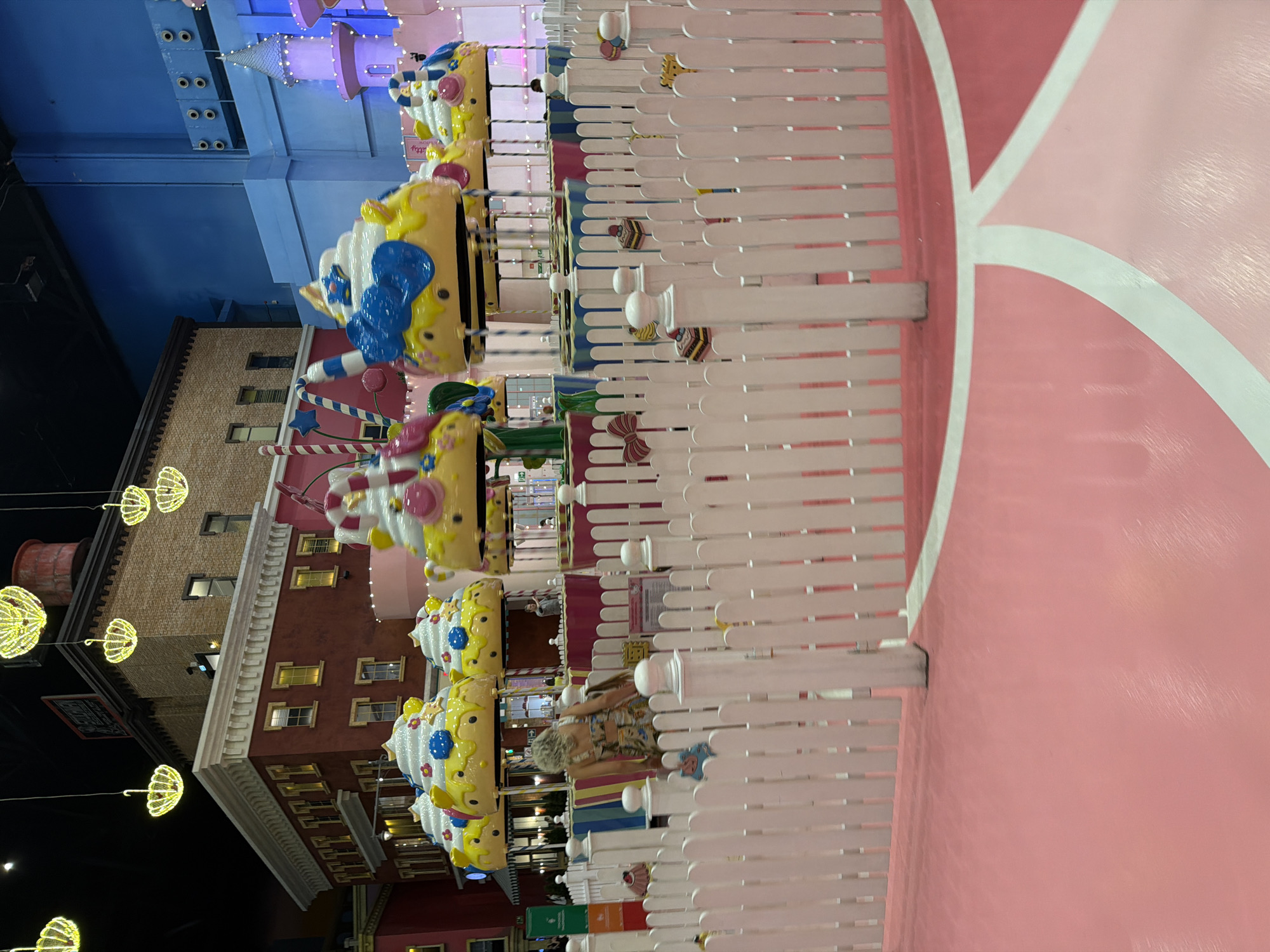The image captures an indoor amusement park area with a carousel ride designed to resemble giant cupcakes. The carousel features multiple cupcake-shaped seats, each decorated with white frosting, colorful candy accents, and yellow bottoms. These seats are suspended from poles. In the foreground, a white picket fence encloses the ride area, with decorative elements such as bows and mini cake replicas attached to the fence. Beyond the fence, the pink floor of the amusement park is visible, marked with white lines creating a patterned design.
In the background, a brick building facade with windows and an ornate roofline is visible. Hanging yellow lights, resembling chandeliers with umbrella-shaped shades, are suspended from the ceiling. Further in the background, a large blue wall with air vents and a pink castle-like structure can be seen, suggesting different themed zones within the park.
One person, a woman with blonde, curly hair, is visible near the fence, leaning forward and appearing to be interacting with something on the ground. Her back is mostly to the camera, and she is wearing a patterned dress. Other individuals are visible in the distance, further back in the amusement park, but they are not distinct enough to identify. The overall environment is brightly lit and appears to be designed for children's entertainment. The location is an indoor amusement park.