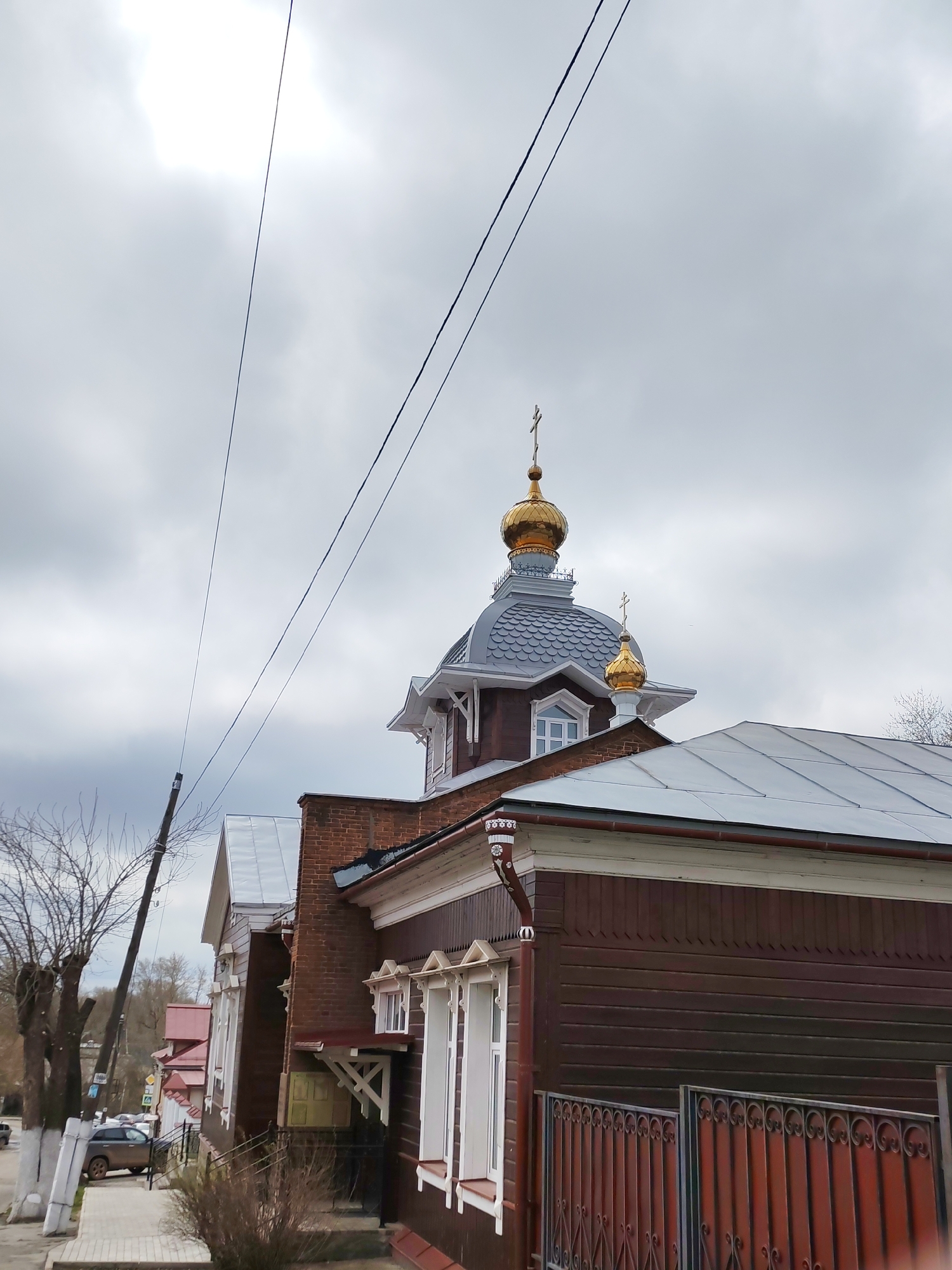 The image shows a view of the Church of the Holy Great Martyr Paraskeva Pyatnitsa, located in Izhevsk, Russia. The church has dark brown wooden siding, white window frames, and a gray roof. A prominent feature is the golden domes topped with crosses. Power lines run across the top left portion of the frame. In the background, there are other buildings and trees, with a gray, overcast sky overhead. A car is parked on the side of the road in the lower left background. A fence runs along the bottom right.