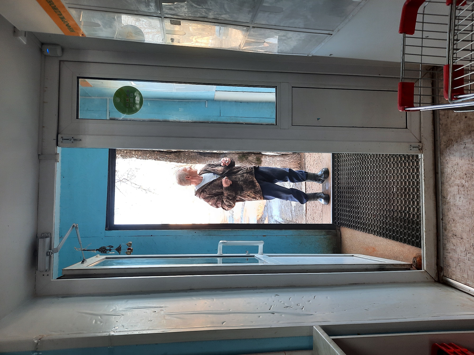 A doorway, seemingly the entrance to a shop, is visible. An elderly man wearing a camo jacket and dark pants stands in the doorway. The shop appears to be in Krasnogorskoye, Russia. The scene includes a doormat and a shopping cart.