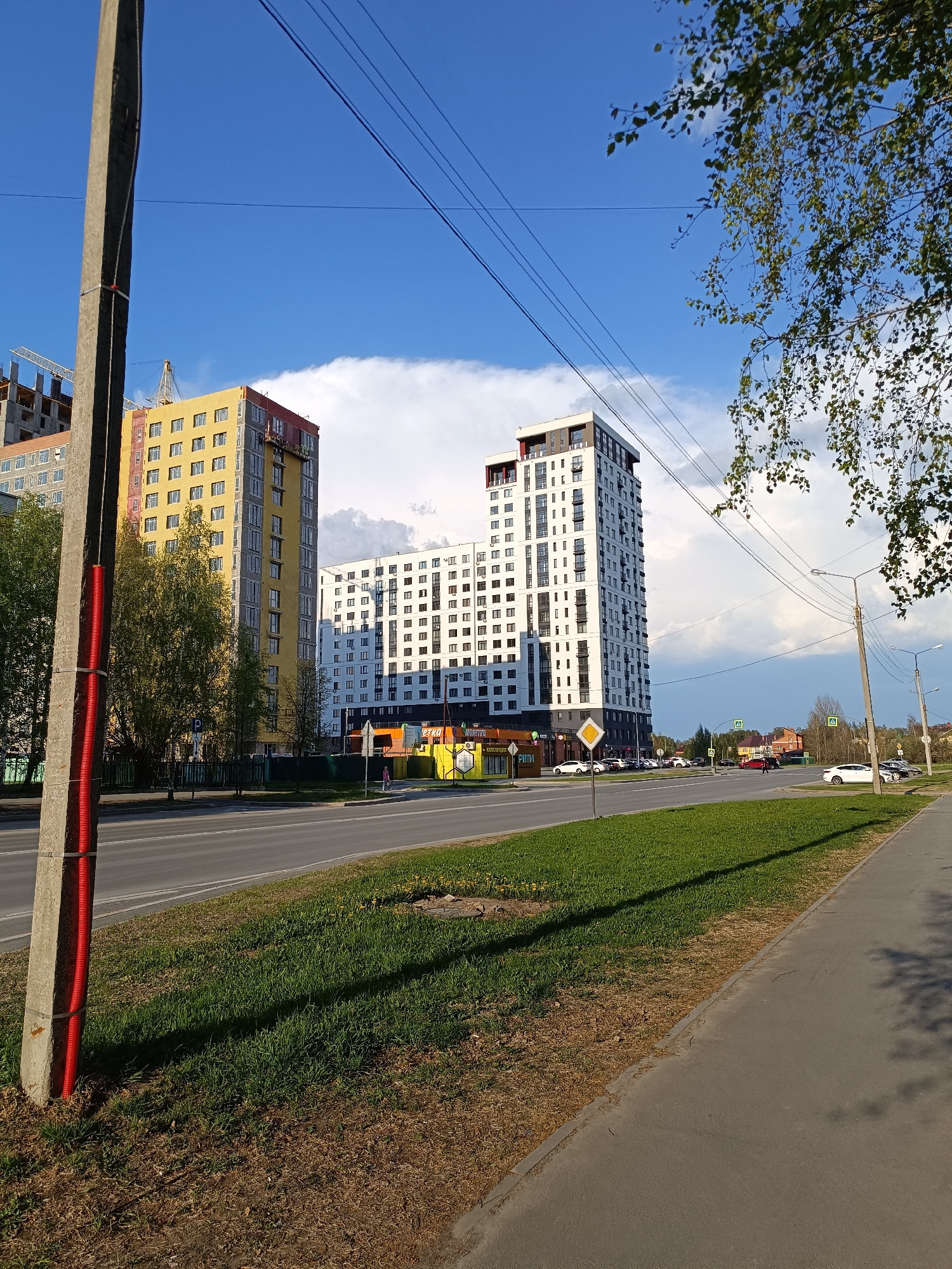 The image depicts a street scene in Tobolsk, Russia, with several tall apartment buildings in the background. The building on the left is under construction with a yellow exterior, the middle building has a white exterior with a brown base. An asphalt road runs through the scene, with a grassy median separating the lanes. Cars are parked on both sides of the road. A sidewalk is visible on the right side of the image. In the foreground, there is a concrete utility pole with a red cable attached, located on a grassy area. Over the street, are power lines, connecting to the pole. The sky is blue with scattered white clouds. Trees with green leaves are present on the right side of the image.