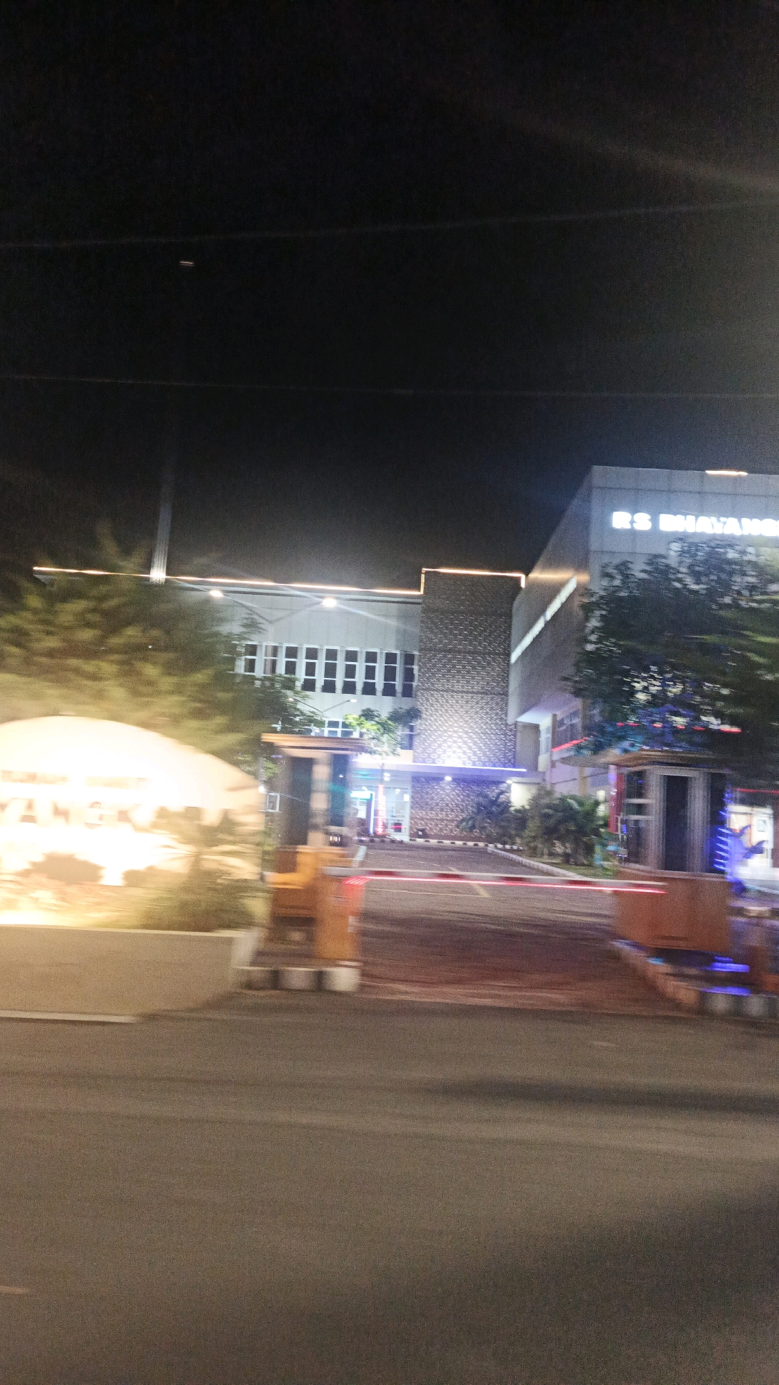 The image is a night-time exterior shot of a modern building with a sign that reads "RS BHAYANGKARA". The building appears to be a hospital. There are trees and foliage visible around the building, and a road with a barrier arm in the foreground, suggesting an entrance or exit to a facility. The lighting is artificial, with illuminated signs and lights from within the building. The overall impression is a well-lit, active location at night in an urban or semi-urban setting.