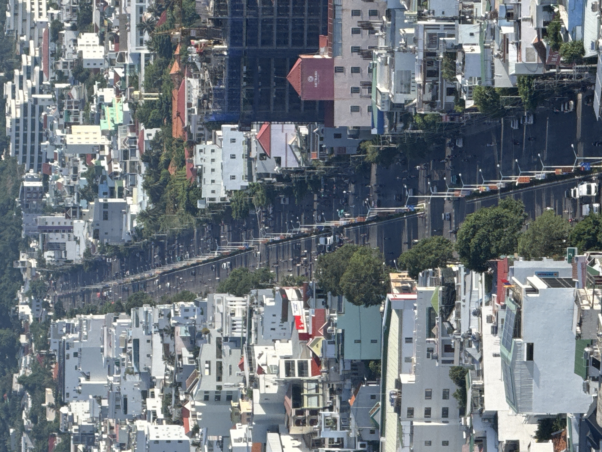 This is an aerial, telephoto view of a city street in Nha Trang, Vietnam. The street, which runs lengthwise through the frame, is lined with numerous buildings, many of which are white or light-colored with red or dark roofs. The buildings vary in height and architectural style, from multi-story apartment blocks to smaller houses. Many of the buildings have balconies and windows visible. Trees and vegetation are interspersed between the buildings and along the sides of the street, with a denser green area visible in the background, possibly a hillside.
The street itself is paved and appears to be a busy thoroughfare. There are several vehicles on the road, including cars, white vans, and numerous motorcycles, indicating active traffic. Streetlights are visible along both sides of the road. Pedestrians are also present on the sidewalks and walking near the road.
Some notable structures include a tall, dark building that appears to be under construction, with blue netting or sheeting covering parts of it. On one of the lighter-colored buildings, a prominent red sign with white lettering, possibly a business name, is visible. Another building has a red, pitched roof and a sign that says "METRANG". Some rooftops have air conditioning units or solar panels. The overall impression is a densely populated urban environment. The lighting suggests it is daytime, possibly late afternoon due to the shadows.