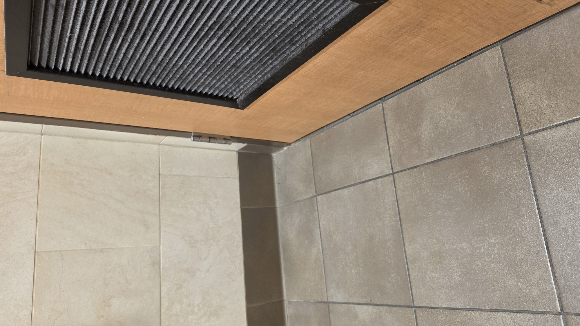 The image shows a close-up view of the corner of a room with tiled walls and floor, and a wooden door with a vent. The wall tiles are beige and rectangular, laid in a horizontal pattern. The floor tiles are square and a light brown or taupe color, with dark grout lines. A dark brown baseboard runs along the bottom of the wall. To the right of the wall is a wooden door, with a darker brown frame around a rectangular vent that has horizontal slats. The door appears to be hinged on the left side from the perspective of the image. There are no people or animals visible in the image. The scene is indoors, and the lighting suggests it is a well-lit interior space. The location of Haughton, United States of America is not visually discernible from the image itself; this information is provided externally.