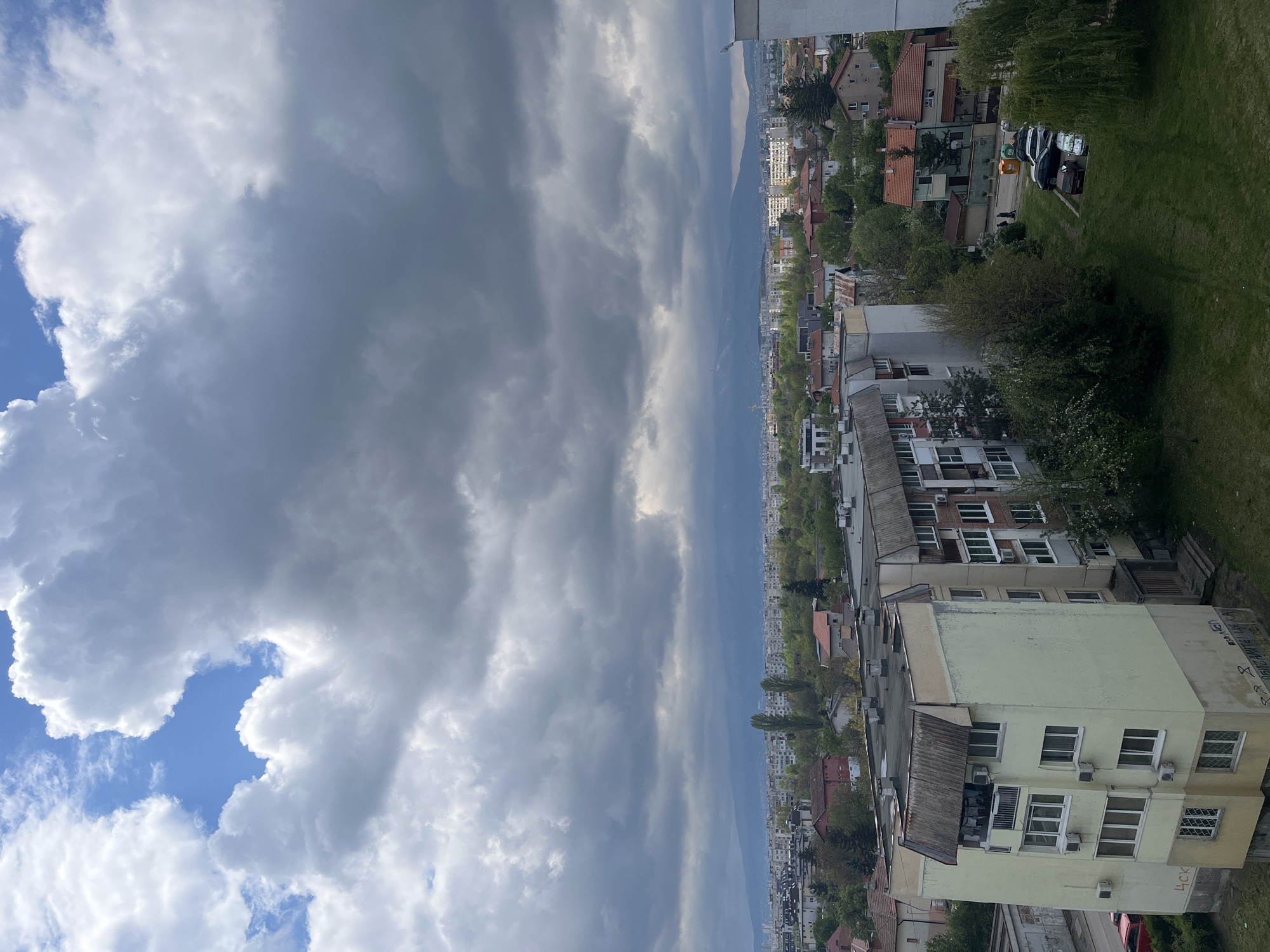 This is an elevated, outdoor view of a neighborhood in Sofia, Bulgaria. The foreground is dominated by light-colored apartment buildings. Further into the middle ground are other residential buildings, houses with red tile roofs, and greenery (trees and grass). In the background, a hazy mountain range is visible along the horizon. Above the buildings, the sky is filled with gray and white storm clouds, with a small patch of blue sky visible in the lower left. A parking area with parked cars and bins is visible. A few people walking on the sidewalk near the parked cars.