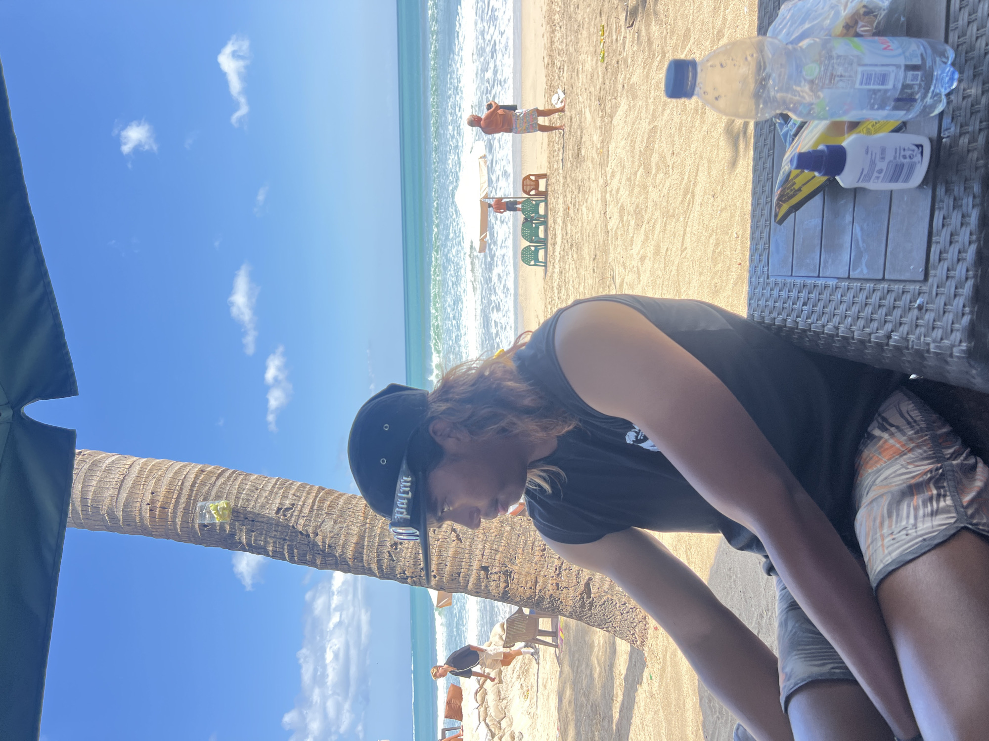 A man wearing a black t-shirt, shorts, and a black baseball cap with a white logo sits on a wicker chair facing left, looking out at a beach scene in Kuta, Indonesia. The man has long, light brown hair. To his left is a tall palm tree with a clear plastic bag containing yellow objects attached to it. In the background, people are visible on the sandy beach. Some are walking, while others are sitting in chairs. A white umbrella shades a section of the beach, and waves are crashing on the shore. A blue plastic water bottle and a small white bottle with a blue cap are on a wicker table in front of the man. The sky is blue with scattered white clouds.