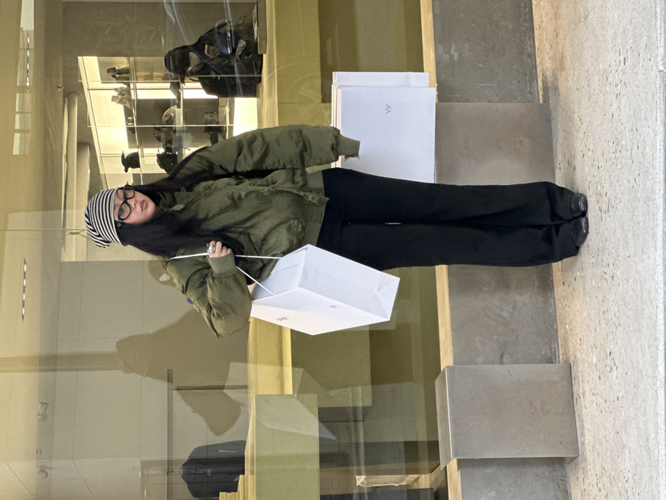 The image presents an East Asian individual standing on a concrete sidewalk in front of a modern retail storefront. The setting is a high-end shopping district in Seoul, South Korea, consistent with areas such as Cheongdam-dong or Apgujeong-ro in Gangnam-gu.
The central figure has long dark hair and wears a black and white striped beanie, black-framed eyeglasses, an olive green bomber jacket, black wide-leg trousers, and black flat shoes. A silver-colored ring is visible on their right hand. They are holding a large, white paper shopping bag with white string handles in front of them, using both hands.
The background consists of a large glass store window reflecting the interior and street elements. Inside the store, visible through the glass and in reflection, are clothing items on hangers, display shelves holding various products, and sections of a yellowish-brown counter. A second individual, wearing a dark baseball cap and jacket, is partially visible in the reflection inside the store, appearing to be observing a device. To the right of the main subject, a similar white paper shopping bag displaying the letters "YY" is placed on a low, light gray concrete ledge. To the left of the main subject, a small, dark rectangular concrete block rests on the sidewalk. The ground beneath the subject is composed of light gray concrete.