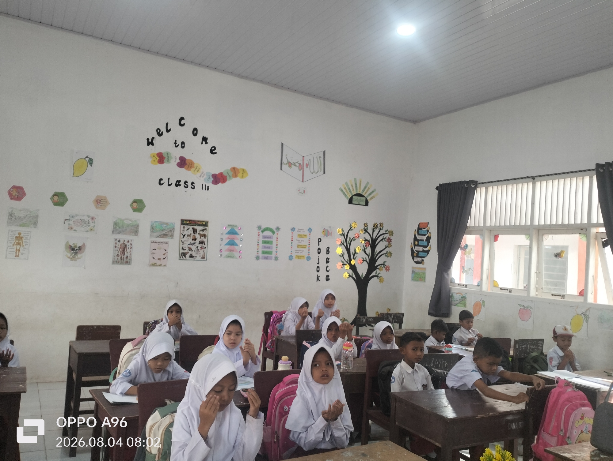 The image shows a classroom of young students in what appears to be a school in Keudekarieng, Indonesia. The students, mostly girls wearing white hijabs and uniforms, are seated at wooden desks. Some students are looking at the camera, while others are engaged in activities at their desks or looking at the front of the classroom. The classroom walls are decorated with various educational posters, including a "Welcome to Class IIB" banner, animal charts, and what appears to be religious calligraphy. Natural light streams in from a large window at the back of the room, which is covered by blinds and gray curtains. The ceiling is paneled and lit by fluorescent lights. A watermark in the bottom left corner indicates the image was taken with an OPPO A96 on August 4, 2026, at 08:02.