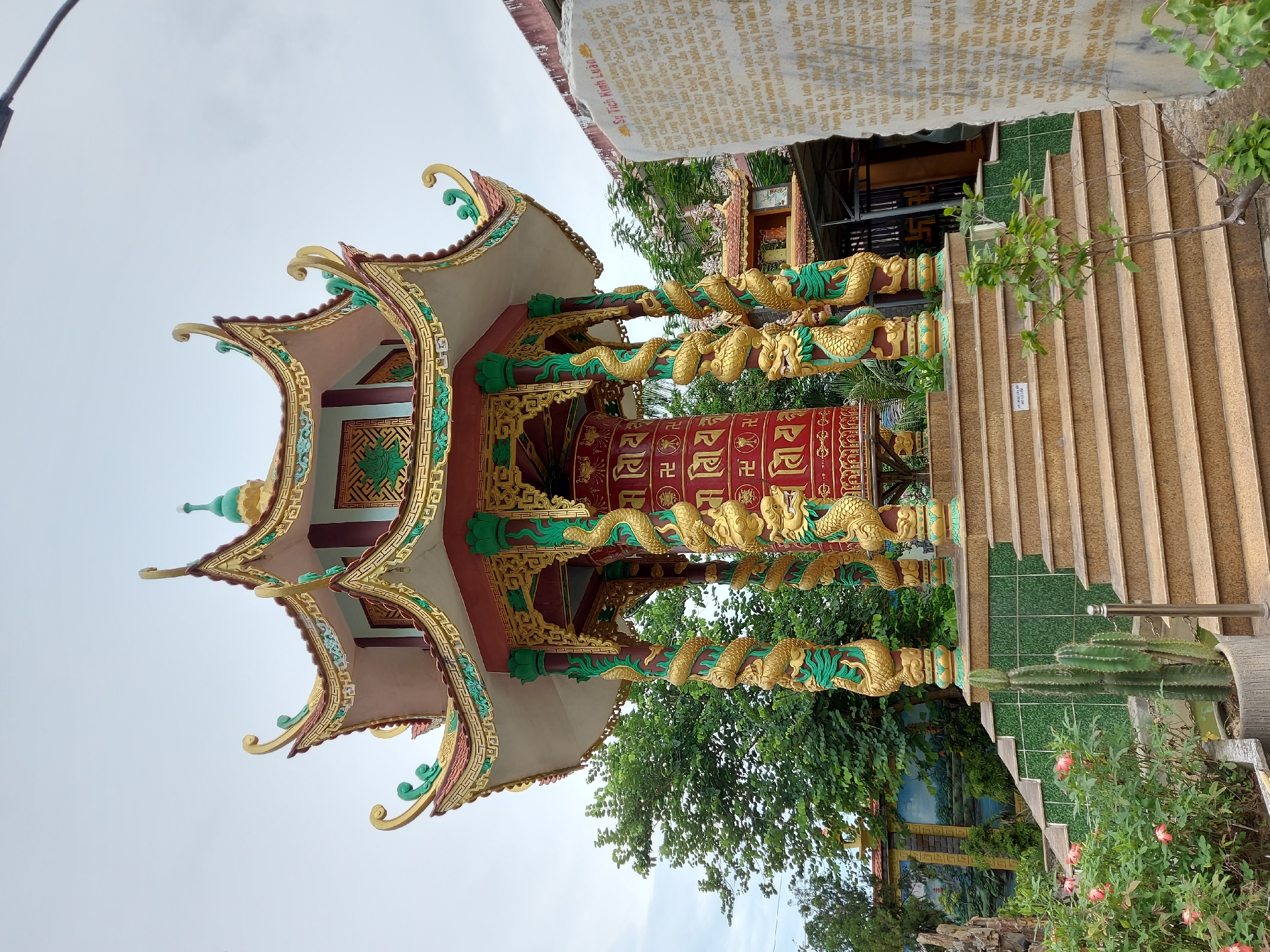 The image depicts a vibrant, ornate Buddhist prayer wheel structure situated in a garden setting. The structure itself is a multi-tiered pavilion with gracefully curved, upturned eaves decorated with gold and green accents. The central element is a large, red, cylindrical prayer wheel adorned with gold Buddhist symbols and Tibetan script. The prayer wheel is supported by four elaborately decorated columns, each featuring a coiled golden dragon wrapped around a green, fluted base.
A wide, light brown staircase with several steps leads up to a platform at the base of the prayer wheel structure. The platform is tiled with green ceramic squares. To the left of the stairs, there is a large planter containing several tall, columnar cacti and some pink flowering bushes. To the right of the stairs, a large, upright stone monument is inscribed with text, presumably religious in nature, in a Vietnamese script.
In the background, lush green trees form a dense canopy. Behind the prayer wheel structure, a smaller building with a similar architectural style can be seen, and further back, a wall decorated with a landscape mural is visible. The sky is overcast with a light gray color. The overall impression is one of spiritual tranquility and artistic craftsmanship within a well-maintained garden. The scene is likely a temple or a Buddhist shrine. While the image itself does not explicitly state the location, such elaborate prayer wheels and architecture are common in Buddhist sites in Southeast Asia, including Vietnam. It is plausible this location is within the city of Đà Nẵng, Vietnam.