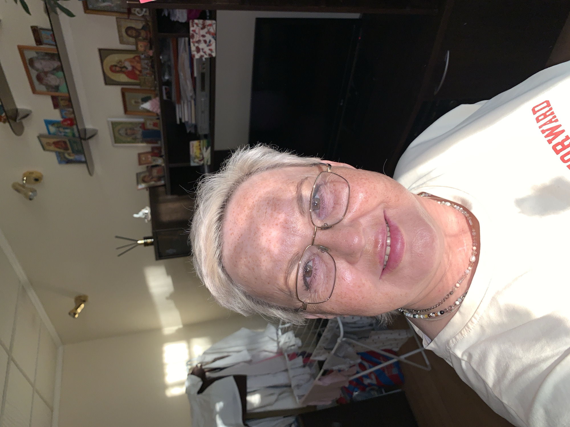 A middle-aged Caucasian woman with short, light blonde hair and fair skin with freckles is taking a selfie in a dimly lit room. She is wearing a light-colored t-shirt with a red word across the front, partially obscured. She has on glasses with gold frames and two necklaces. The background shows a shelf filled with framed pictures, some of which appear to be religious icons. There is also a television set and a clothes drying rack with laundry. The room is illuminated by natural light entering from a window on the left, casting a shadow on the wall and ceiling. The location is Chaikovsky, Russia.