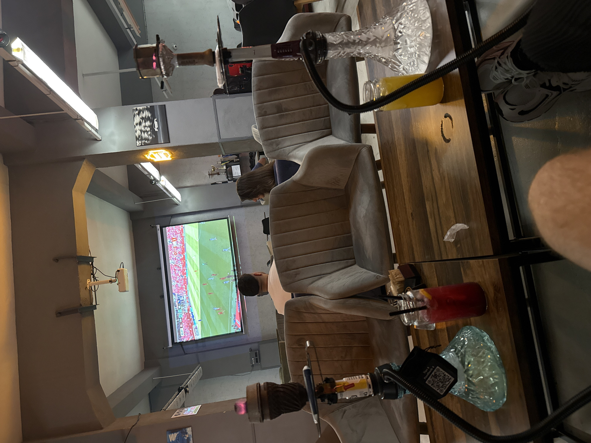 The image depicts the interior of a modern lounge or cafe in Plovdiv, Bulgaria, where a football match is being projected onto a large screen. The room features comfortable, plush seating and a dark wood table in the foreground. Two shisha pipes are prominently displayed, one on the table and another on the right side of the image, suggesting the venue's primary function. On the table, a red cocktail with straws and a glass bottle of orange juice are visible. The overall ambiance is relaxed and social, with patrons engaged in watching the sporting event. The lighting appears to be a mix of ambient and artificial, with neon signs and overhead fluorescent lights contributing to the atmosphere. The presence of trophies on a shelf in the background hints at a potential theme or a history of competitive events hosted at the venue.