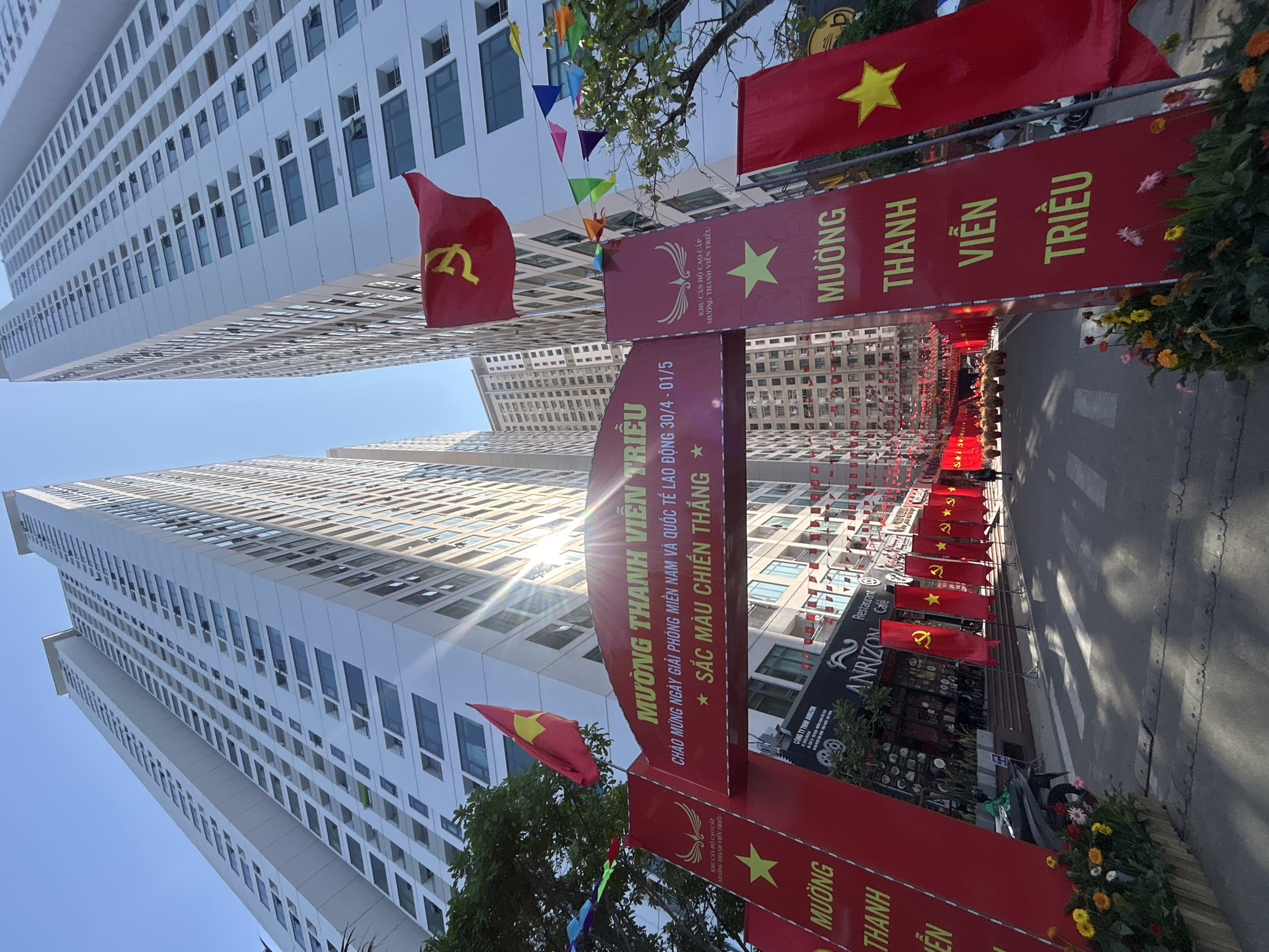This vertical-oriented photo captures a festive streetscape in Bắc Nha Trang, Vietnam, adorned with red banners and flags under a bright, clear blue sky. Tall, modern white apartment buildings, identified as the "MƯỜNG THANH VIỄN TRIỀU" high-end complex, line both sides of the road, reflecting the intense sunlight.
The street is decorated for a celebratory event. Large red banners span the pathway, forming an archway, welcoming "NGÀY GIẢI PHÓNG MIỀN NAM VÀ QUỐC TẾ LAO ĐỘNG 30/4 - 01/5" (Day of Liberation of Southern Vietnam and International Labor Day 30/4 - 01/5), accompanied by the phrase "* SẮC MÀU CHIẾN THẮNG *" (Colors of Victory). Smaller red flags featuring the yellow star of Vietnam and the hammer and sickle symbol of the Communist Party of Vietnam alternate along the sidewalk. Colorful triangular pennants are strung across the street, enhancing the celebratory mood.
In the background, a few indistinct figures can be seen walking. On the right side, a storefront for "ANRIZON Restaurant Café" is visible. Flower beds with various yellow, pink, and white blossoms add natural beauty to the urban setting. The bright sunshine, causing a lens flare in the center, indicates a clear and sunny daytime. The scene conveys a strong sense of national pride and celebration in an urban Vietnamese environment.