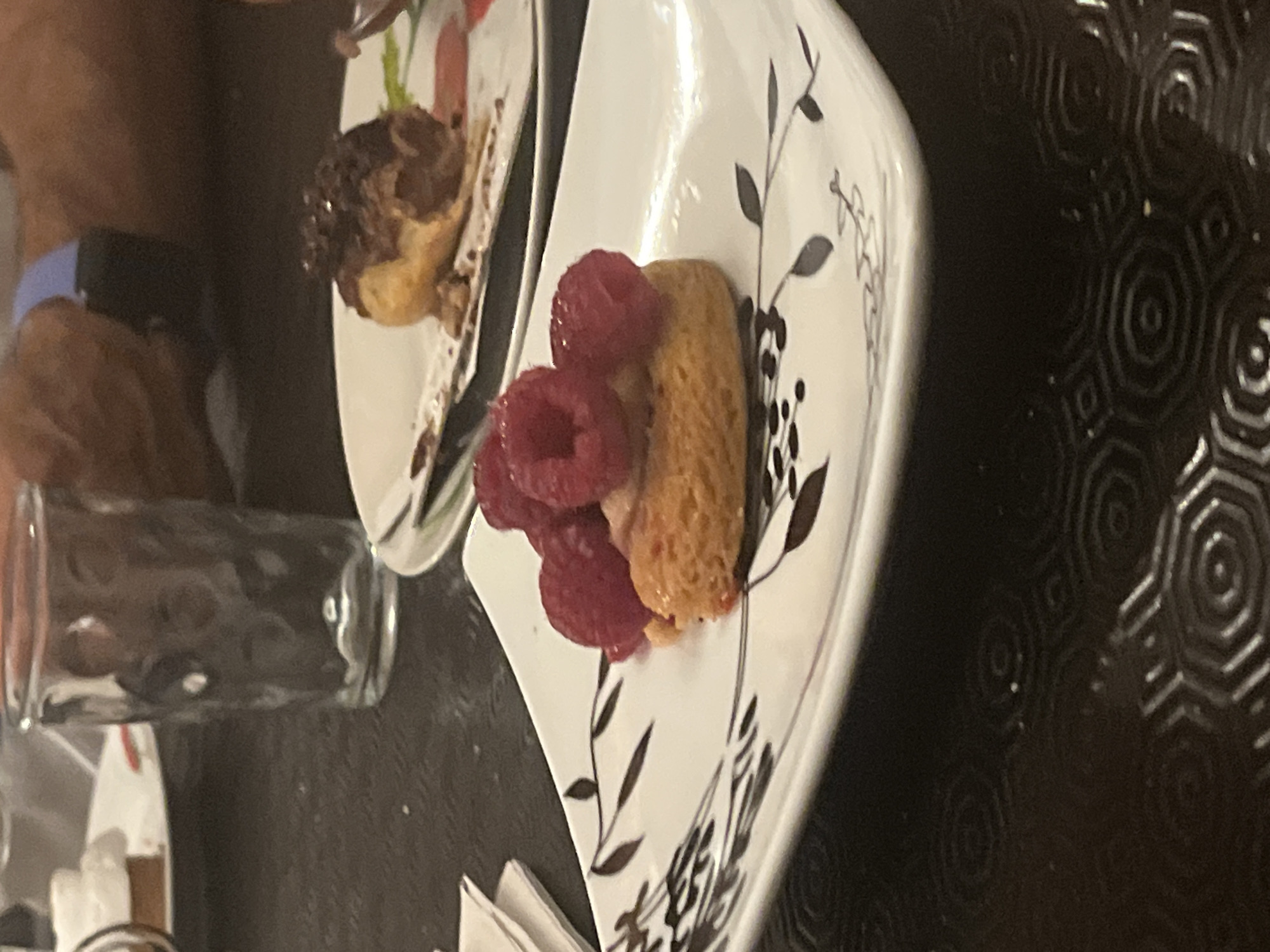The image displays a close-up view of a dessert with raspberries on a decorative white plate. The plate has a dark, leafy pattern on its edge. In the background, another dessert, possibly a chocolate éclair, is visible on a white plate with a red garnish. A clear glass with ripples is positioned to the left of the desserts. The table surface appears to be dark and textured. The scene is likely set in a restaurant or dining establishment, possibly in Lorette, France, given the provided context, though this cannot be definitively determined from the image itself.