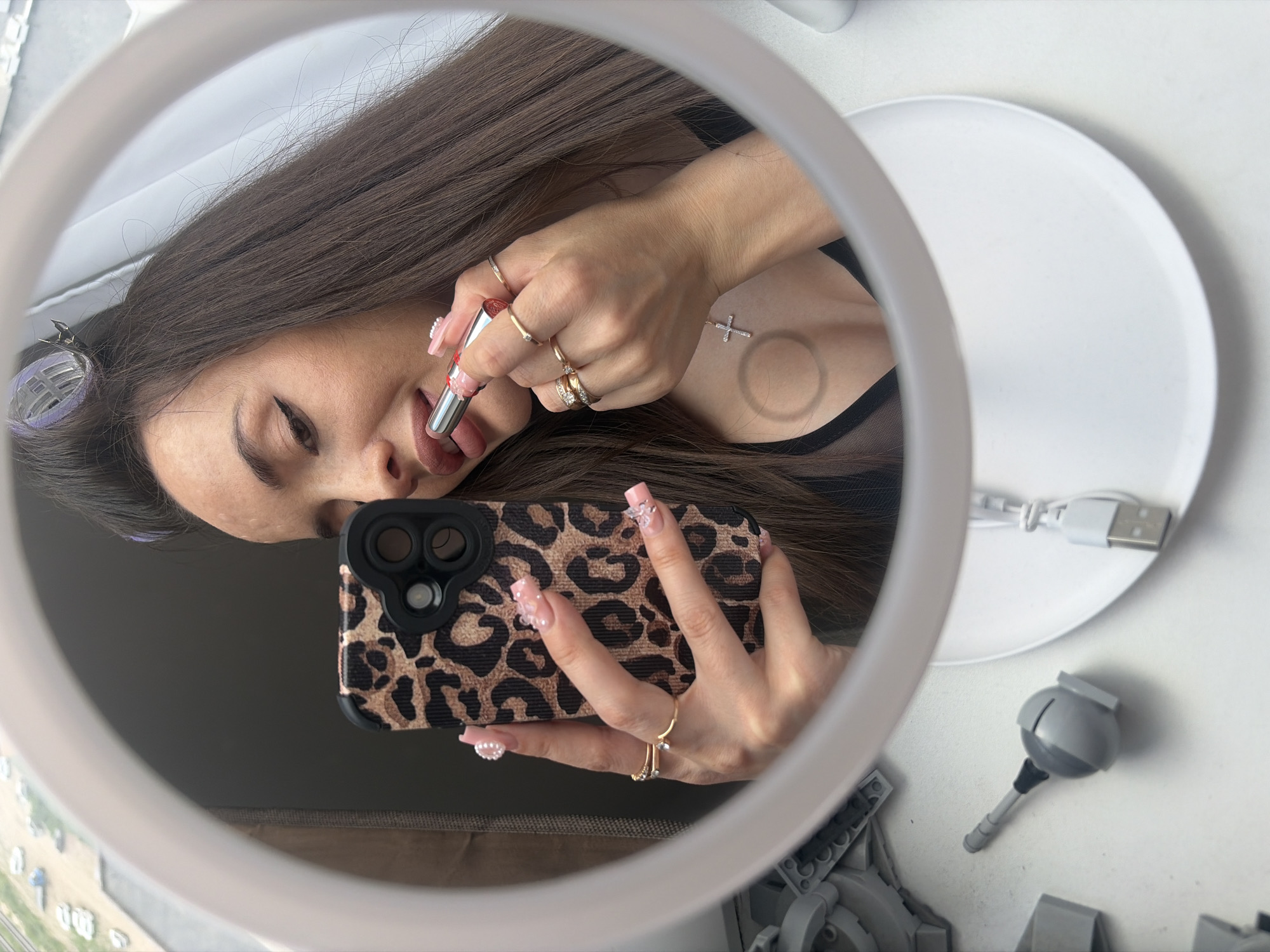 The image depicts a woman taking a mirror selfie indoors, in what is described as the city of Saint Petersburg, Russia. The woman, with long dark brown hair styled with two visible purple hair rollers, is reflected from the chest up in a circular mirror framed by a prominent white ring light. Her right hand is positioned at her mouth, applying a silver-colored lipstick with red accents to her lips. Her left hand holds a smartphone with a leopard print case, directed towards the mirror to capture the reflection.
Both of the woman's hands are adorned with multiple gold-toned rings, some featuring small clear stones, on various fingers. Her fingernails are manicured with pink polish and elaborate 3D nail art, including pearls on the thumb and ring finger of the left hand, and small bows or rhinestones on the index finger of the left hand and the ring finger of the right hand. She is wearing a black top, possibly sheer on the upper chest area, and a delicate chain with a silver-colored cross pendant around her neck.
The mirror's base rests on a white surface, likely a table or desk. To the left of the mirror, several gray Lego or similar building block pieces are visible, including one piece resembling a spherical joint with a long rod extending from it. To the right, a white coiled USB-A charging cable is placed on a shallow white plate or tray. Through a window reflected in the upper left of the mirror, an urban outdoor scene is visible, showing parked white cars and multi-story buildings under an overcast sky, consistent with a city environment.