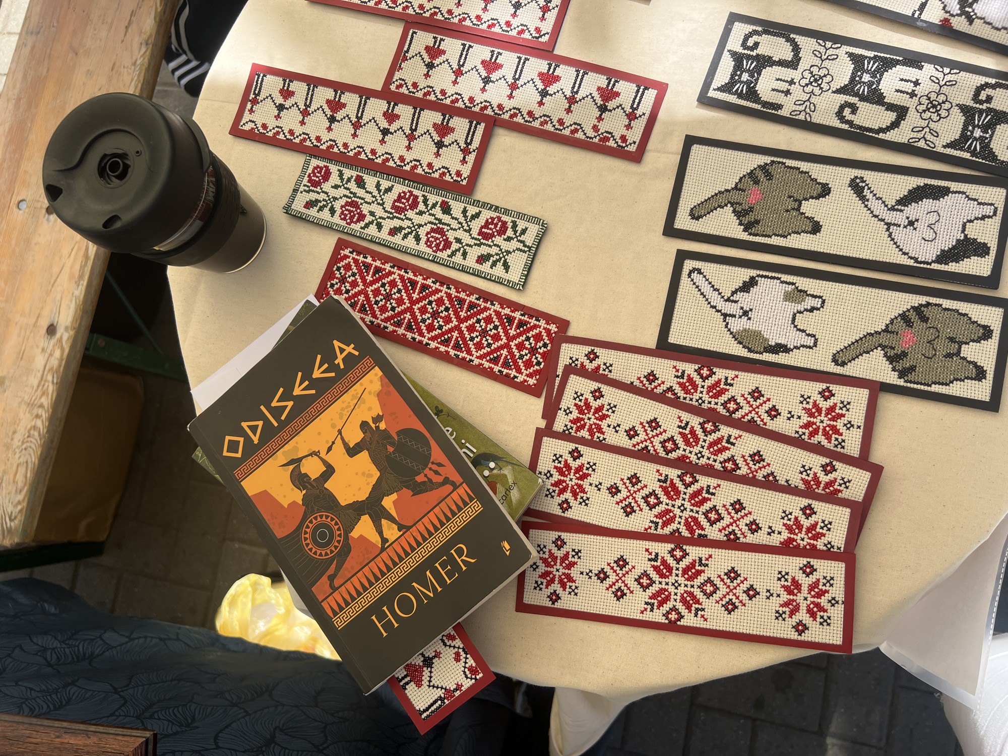 This is a close-up, eye-level, outdoor shot of a table covered with a light-colored cloth. On the table, there is a book titled "ODYSSEY" by HOMER, with an illustration of two warriors on the cover. Scattered around the book are several embroidered bookmarks. Some of these bookmarks feature floral patterns, specifically red roses, while others display geometric designs in red and white. Further down the table, there are bookmarks with pixelated designs of cat bottoms, and another set of bookmarks with black and white cats. In the upper right portion of the image, a black thermal mug with a silver band is visible, resting on the edge of the table. The background shows a wooden bench and a cobblestone ground. The overall scene suggests a market stall or a craft fair. There is no visual information to place this scene specifically in Chișinău, Moldova.