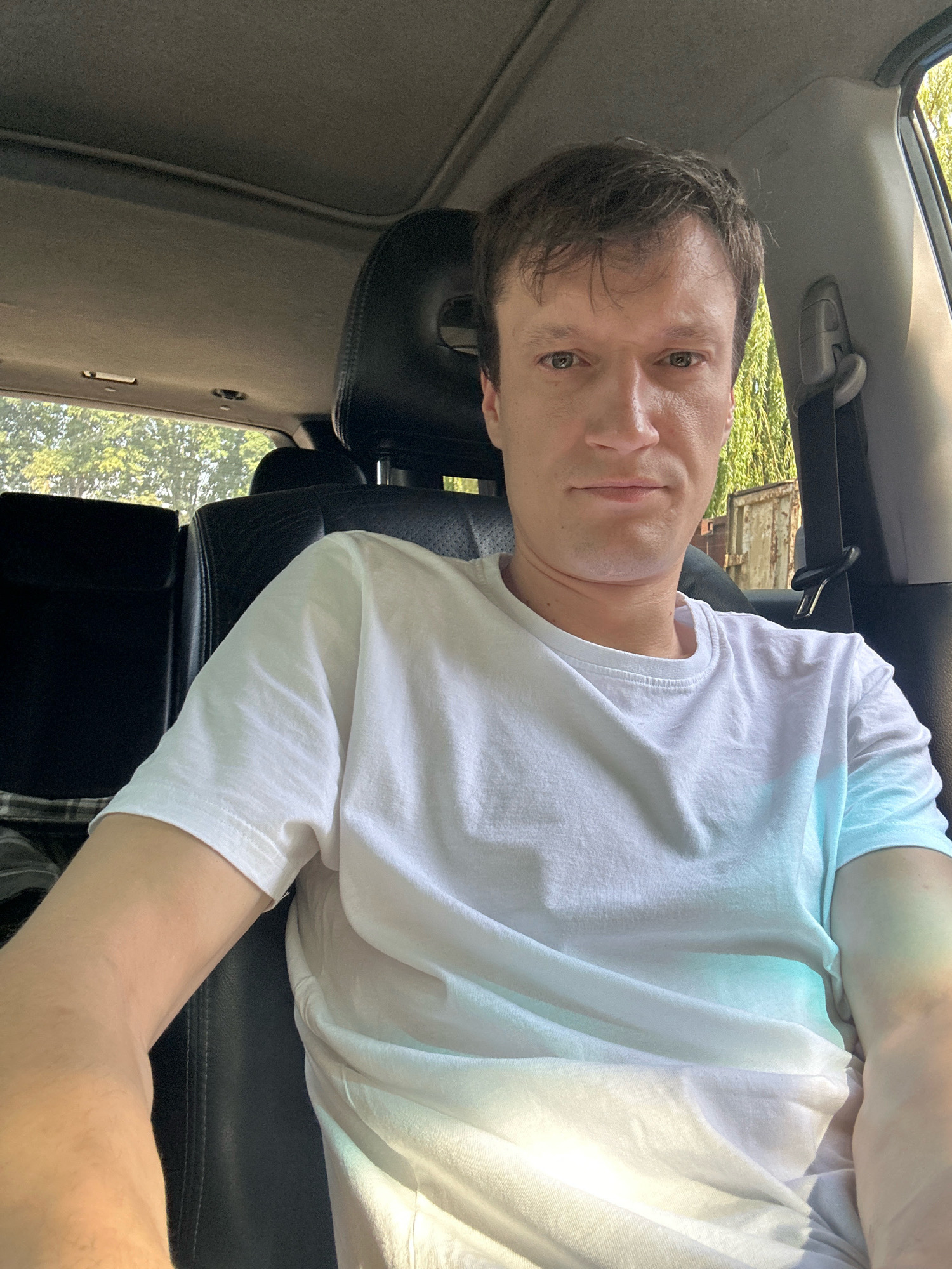 A light-skinned man with short brown hair and green eyes is sitting in the driver's seat of a car, taking a selfie. He is wearing a plain white t-shirt. The interior of the car is visible, including black leather seats, a sunroof, and a seatbelt. Outside the car, trees and a metal structure are visible. The lighting suggests it is daytime. The image was taken in Kamianets-Podilskyi, Ukraine.