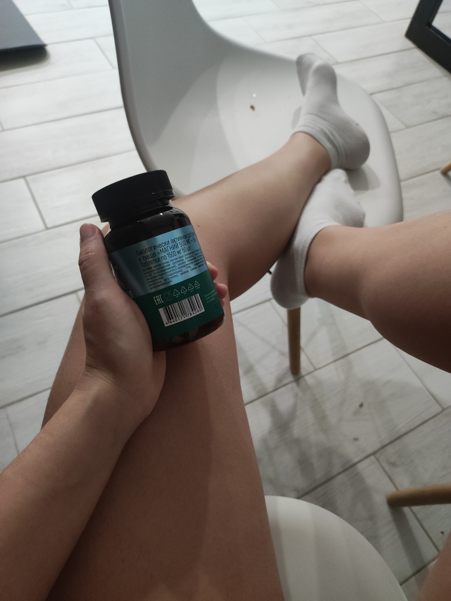 The image depicts a person sitting on a white chair in Votkinsk, Russia, with their legs crossed. They are wearing white socks. In their left hand, they hold a dark green bottle with a black cap. The bottle has text written in Russian, appearing to be a supplement or medication label. The background shows a tiled floor, other parts of the chair, and a glimpse of a wall.