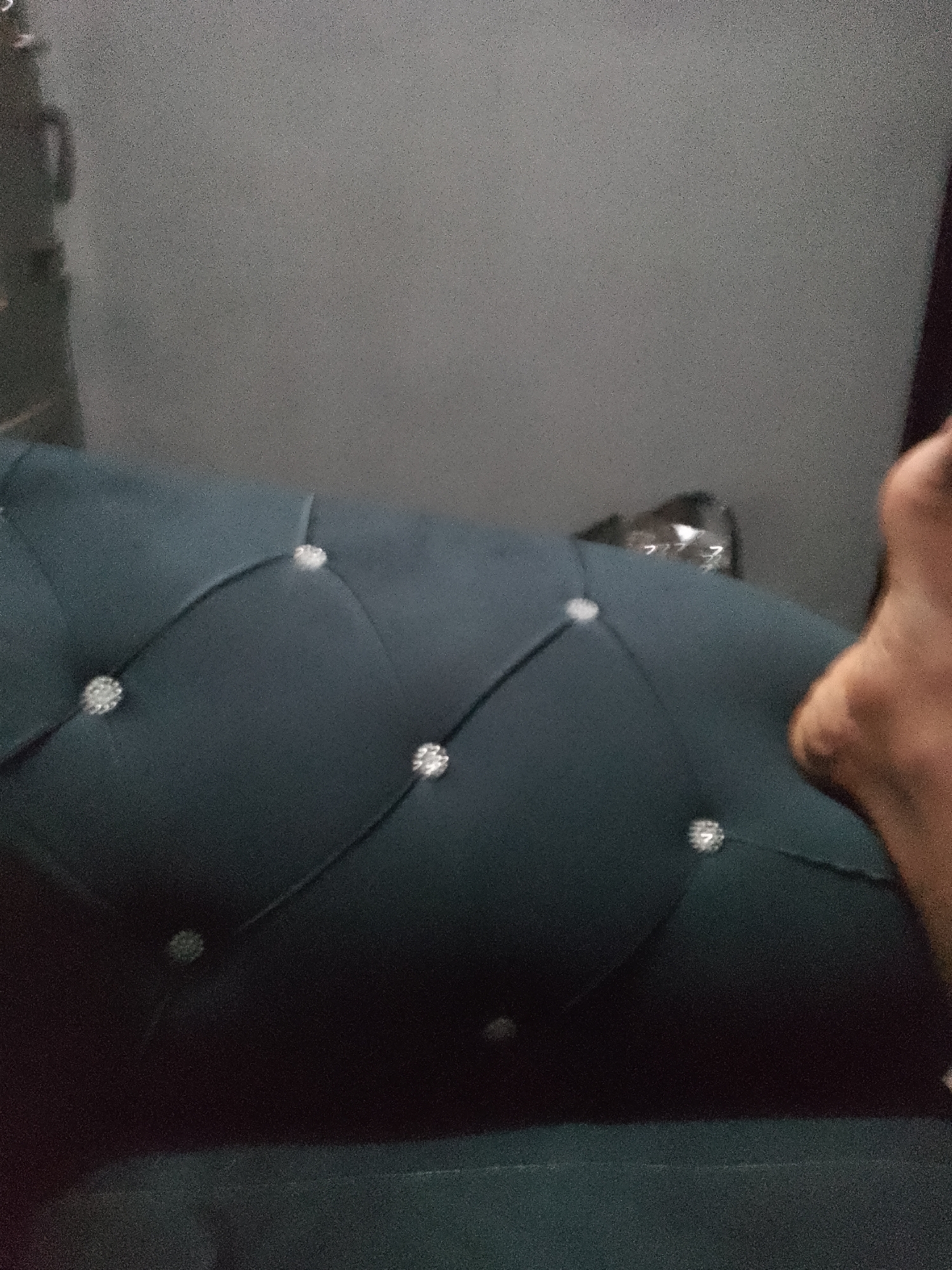 The image shows a close-up of a dark teal tufted upholstery with diamond-shaped patterns created by tufts and accents. The texture of the fabric appears plush. A person's bare foot, with visible skin tone variations and a heel, is positioned on the right side of the frame, partially obscuring the upholstery. The background is out of focus, showing a gray, textured surface, possibly a wall, with a faint reflection of what might be a shelving unit or cabinet on the left. The lighting in the image is dim, contributing to the grainy texture of the photograph. The scene is static, with no discernible action taking place.