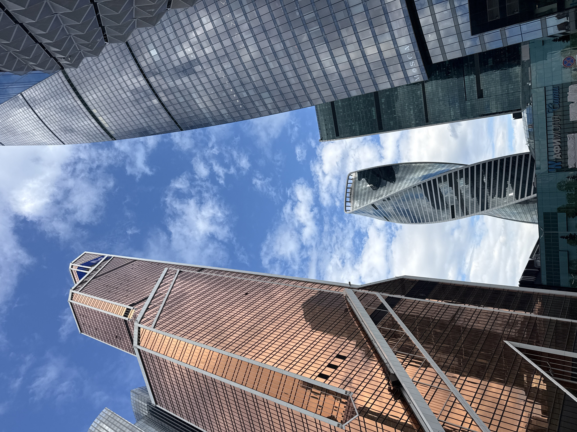 The image shows a low-angle view of several skyscrapers against a blue sky with scattered white clouds. The buildings are modern glass structures with varying designs, including rectangular and twisted shapes. The exterior finishes include copper-colored and silver-toned panels, creating a grid-like pattern. The scene appears to be taken in an urban area, possibly a financial or business district. The angle emphasizes the height and scale of the buildings. There is no visible evidence to suggest the location is in the city of Khimki, Russia.