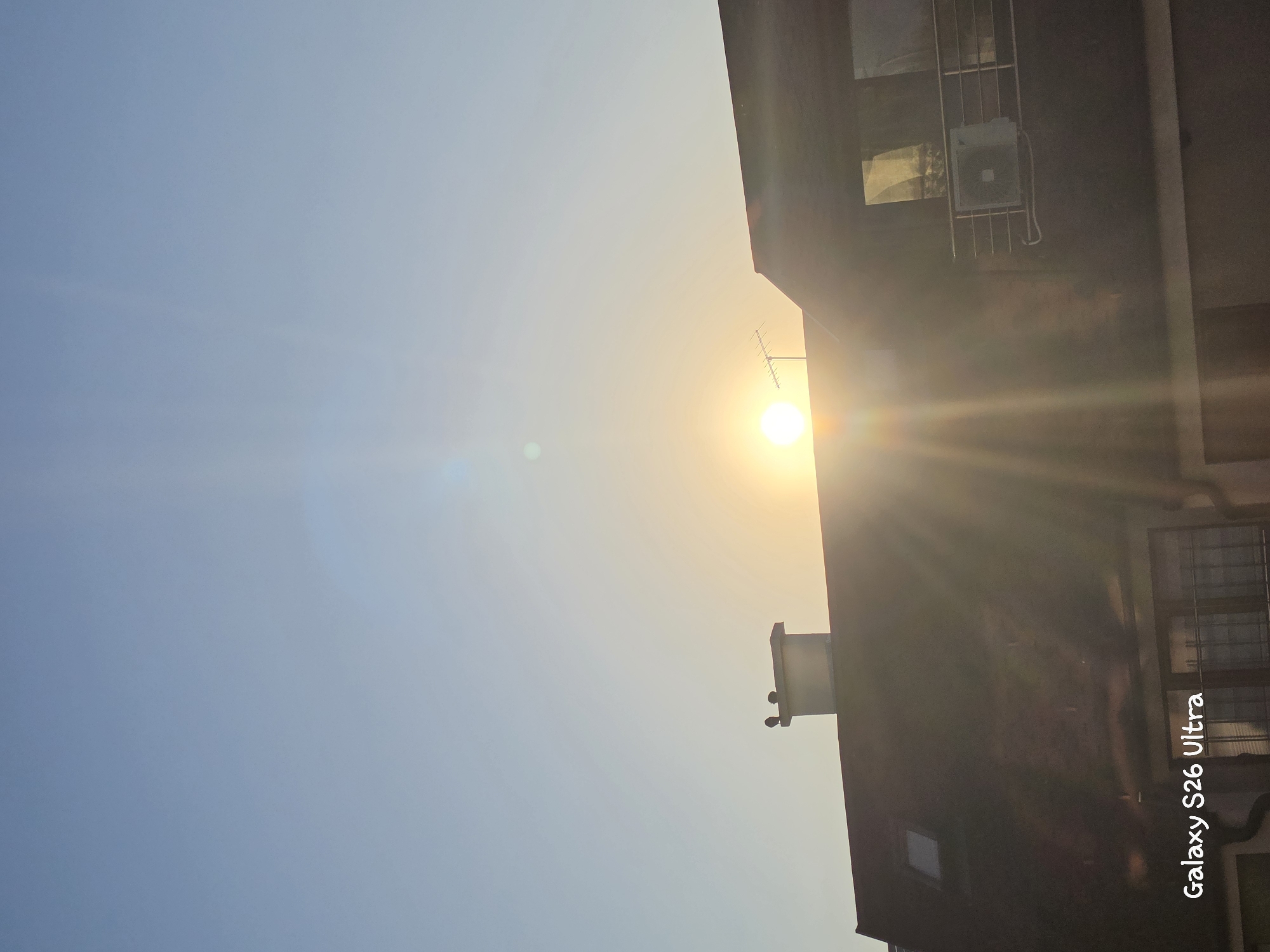 The image depicts a sunset or sunrise over a residential building in Plovdiv, Bulgaria. The sun, a bright orb with visible rays, is positioned near the center of the frame, partially obscured by the roofline of a dark-colored building. A chimney with two birds perched on top is visible on the left side of the roof. An antenna is also visible above the roofline to the right of the chimney. The sky is a clear gradient of light blue, transitioning to a pale yellow near the horizon. The building features a dark, sloped roof with a skylight. A balcony with a metal railing and an air conditioning unit is present on the right side of the building. The bottom left corner of the image contains a watermark reading "Galaxy S26 Ultra". The scene is bathed in warm, golden light characteristic of a low sun angle.
