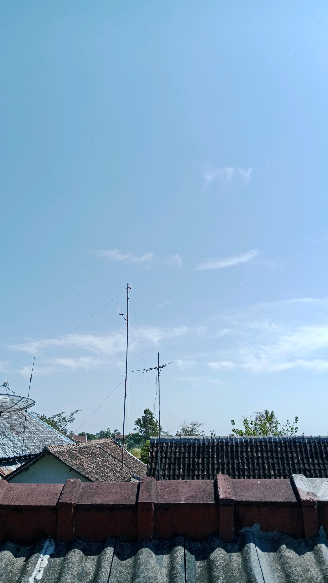 The image depicts a rooftop view of residential buildings in Kebonagung, Indonesia, under a clear blue sky with scattered wispy clouds. In the foreground, the top of two tiled roofs are visible, the closest one a dark gray corrugated material, and the one behind it a red-brown tile with a weathered appearance. Behind these, several other tiled roofs are visible, some with satellite dishes. Two tall antennas with wires extending between them stand out against the sky. Trees and other buildings are visible in the background, suggesting a suburban or urban residential area. The scene is devoid of people.