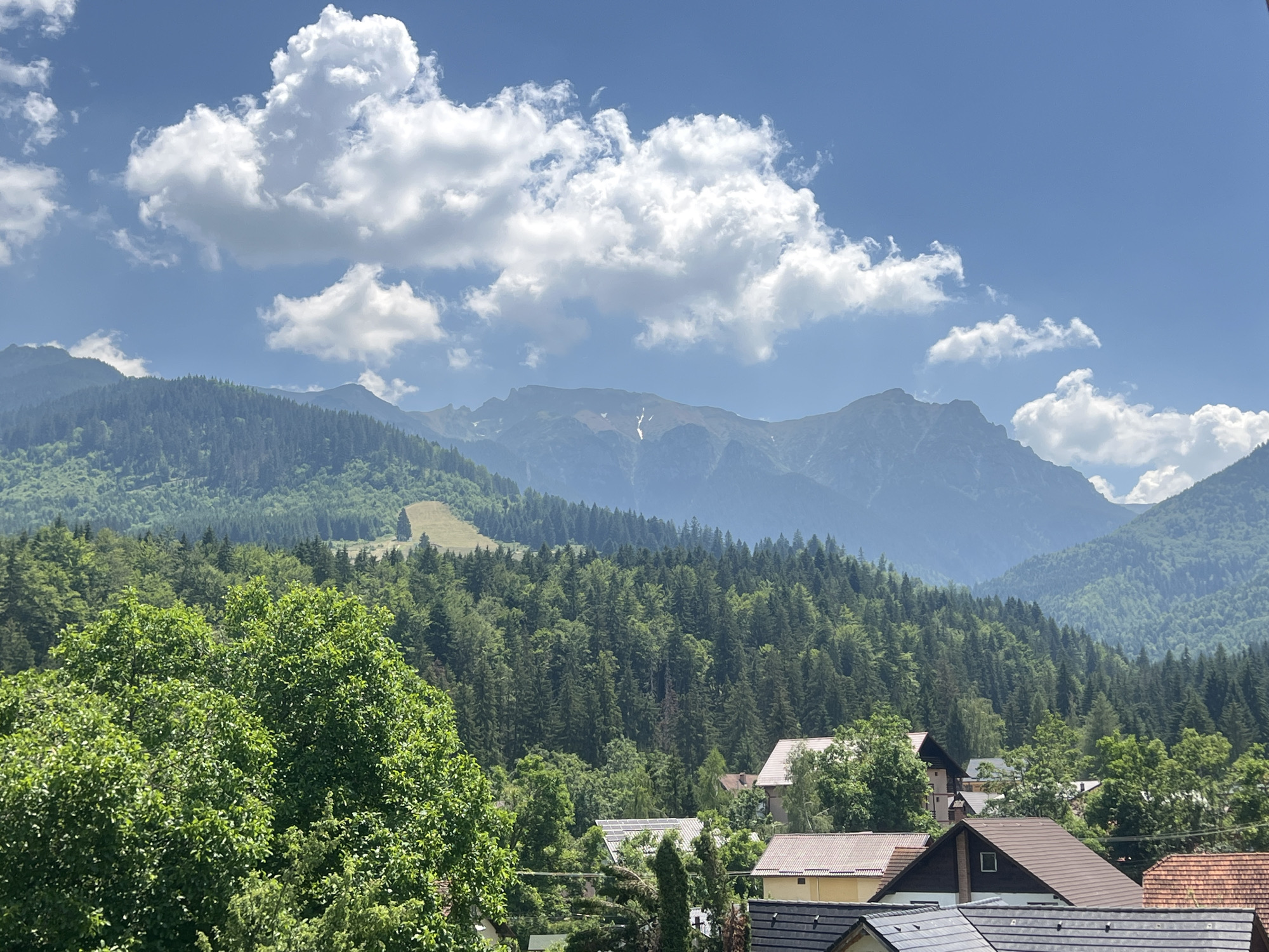 This image depicts a mountain landscape with a village nestled in the foothills. The foreground features lush green trees and the rooftops of several houses with terracotta and dark grey tiles. Some houses have solar panels on their roofs. Beyond the village, rolling hills covered in dense evergreen forests lead up to majestic, jagged mountains in the background. The sky is a clear blue, dotted with fluffy white clouds. The overall scene conveys a sense of natural beauty and tranquility.