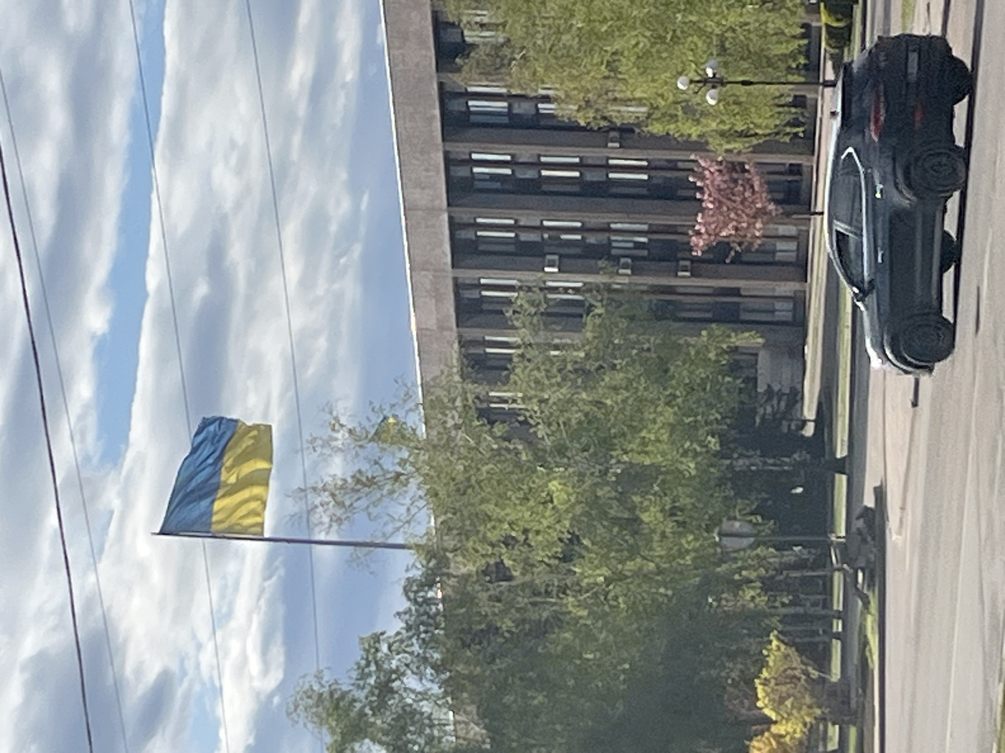 The image shows a view in an urban setting, likely in Unknown, Unknown. The Ukrainian flag is prominently displayed against a cloudy sky. In the background, there is a large, multi-story building with repetitive window patterns. A tree obscures parts of the building and the flag. On the right side of the image, there is a street with a car parked along the curb. A street light is also visible.