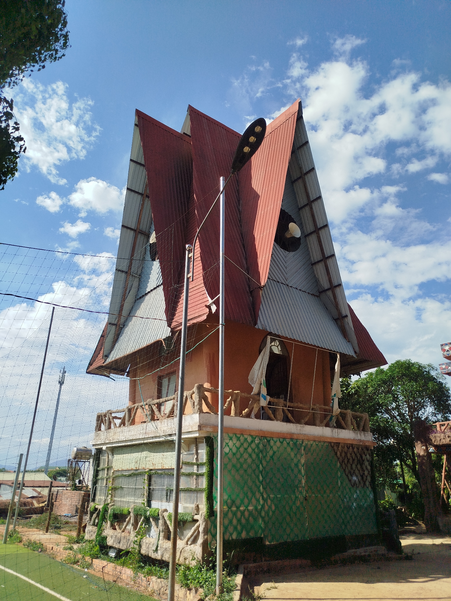 A multi-story building with a distinctive roof stands in An Phu Ward, Vietnam, under a partly cloudy sky. The building features a layered roof design with red and gray corrugated metal. The building is orange and includes a balcony with faux-stone railings. The base of the building has green, diamond-patterned panels. A tall metal light pole stands in front of the building. To the left is a green field enclosed by a net fence. Other buildings and trees are visible in the background.
