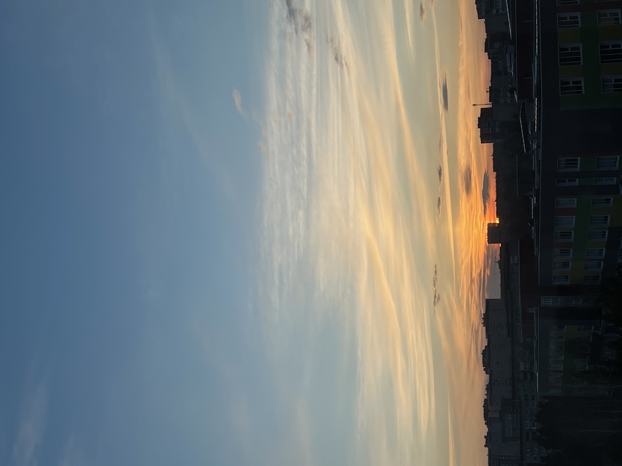 The image depicts a vibrant sunset or sunrise over an urban landscape, consistent with a city like Izhevsk, Russia. The sky dominates the upper portion of the frame, displaying a striking gradient of colors. On the left, it transitions from a clear, calm blue to warm hues of yellow, orange, and soft pink towards the right, where the sun is located. Long, wispy, and streaky clouds extend horizontally across the sky, reflecting the sun's warm light and adding texture to the expansive view. Some darker, more defined cloud patches are visible lower down, silhouetted against the bright horizon.
Below the colorful sky, a silhouette of a city skyline stretches across the lower part of the frame. Numerous buildings, likely apartment blocks and other urban structures, form a dark, jagged line against the brilliant backdrop. The sun, a bright orange glow, is partially visible just above the distant buildings, acting as a radiant focal point. Closer buildings on the far right side of the frame offer more visible detail, including multiple windows and the outlines of their structures. One prominent building on the right features distinct, colorful facade panels in shades of green, yellow, and orange. The perspective suggests the photograph was taken from an elevated position, looking out over the city. The overall scene conveys a serene and beautiful atmosphere at the transition of day. No visible text is present in the image.