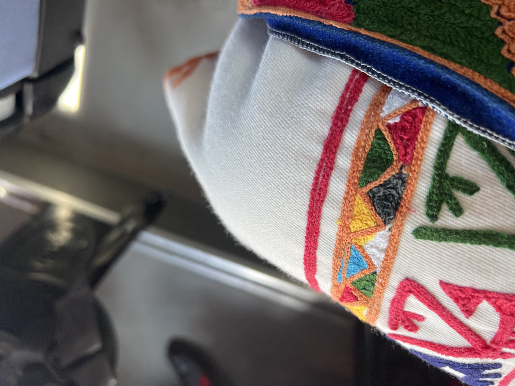 A close-up, high-angle view captures a segment of white textile fabric adorned with vibrant, multi-colored geometric embroidery. The fabric exhibits a visible diagonal twill weave. The embroidery prominently features a horizontal band composed of small, interlocked triangles in red, blue, green, yellow, black, and white threads, delineated by an orange thread border. Below this band, larger, stylized motifs in red, green, and blue threads are directly embroidered onto the white textile. To the right, an attached trim is visible, characterized by a blue, velvet-like band edged with a silver-grey stitch, adjacent to a green embroidered section further outlined in orange thread. The background is significantly out of focus, revealing dark, indistinct shapes on the upper left, suggestive of interior vehicle components, and lighter, equally blurred shapes on the upper right, indicating an illuminated area or window. A dark, abstract form with a red accent is vaguely discernible in the far lower-left quadrant. The scene is located in the city of Slivovo, Bulgaria.