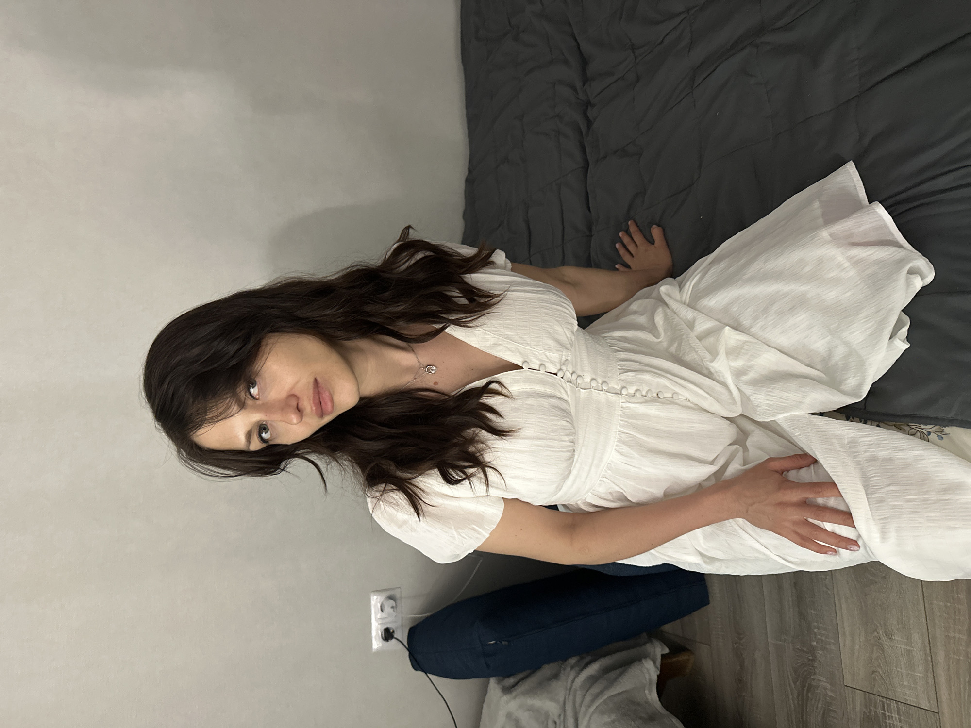 A young woman with dark hair is lying on a bed with a gray comforter. She is wearing a white dress. Her head is near a gray wall with an electrical outlet. A dark blue pillow rests underneath her. The room appears to be indoors, likely in a residential setting, and the flooring is a light-colored wood-grain pattern. The location is stated to be Murino, Russia.