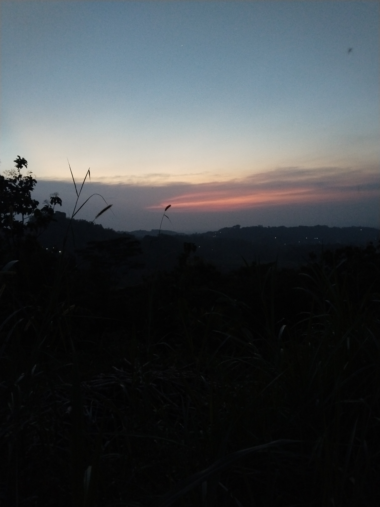 The image depicts a landscape at dusk. The sky transitions from blue to orange and pink at the horizon, indicating a setting or rising sun. In the foreground, dark silhouettes of vegetation, possibly tall grasses and trees, obscure much of the view. Beyond the immediate foreground, a series of dark, undulating hills are visible, with small points of light scattered across them, suggesting a populated area or structures with lights. The location is Unknown, Unknown.