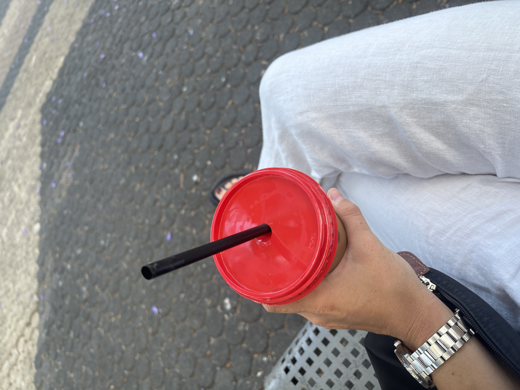 The image shows a close-up view of a person's hand holding a red-lidded cup with a black straw. The person is wearing a silver watch on their left wrist and is dressed in white linen pants. The background consists of a cobblestone-like surface, possibly a sidewalk or plaza, with some shadows cast across it. There are small purple specks scattered on the ground, which could be fallen flower petals. The scene suggests an outdoor setting, likely in a city given the pavement and what appears to be a bench on the left side of the frame. While the exact location in Tel-Aviv is not identifiable from the visual cues alone, the overall ambiance points to a public outdoor space in the city.